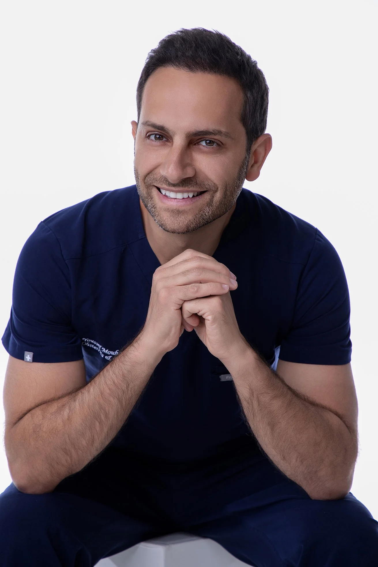 A smiling man with short dark hair, beard, and mustache, wearing a navy blue medical uniform, sitting with hands clasped.