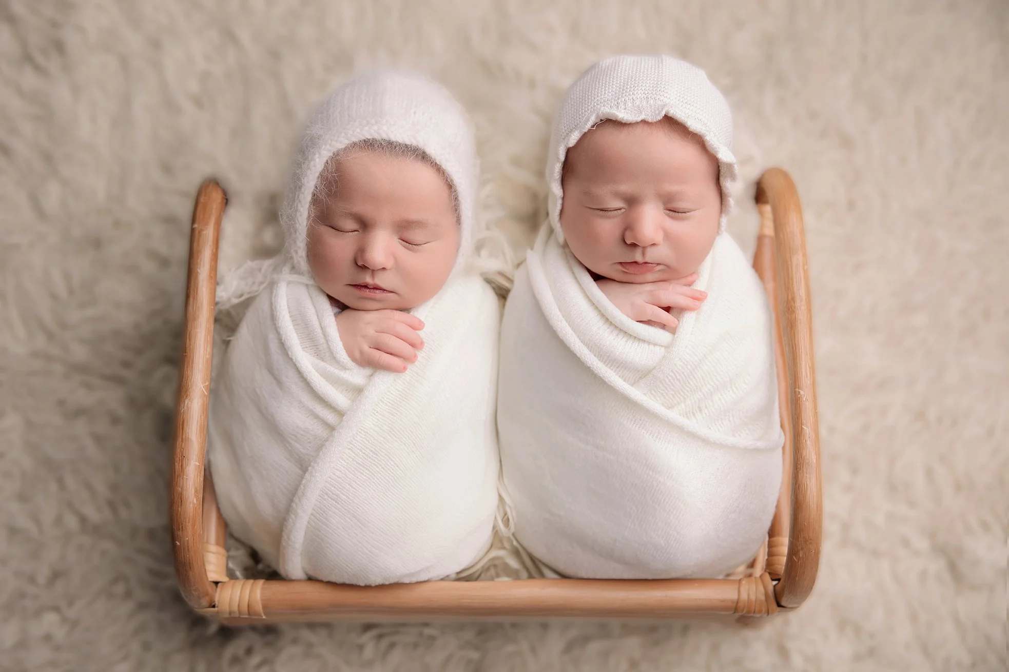 Two babies sleeping wrapped in white blankets, lying side by side in a wicker basket, wearing white knit hats.