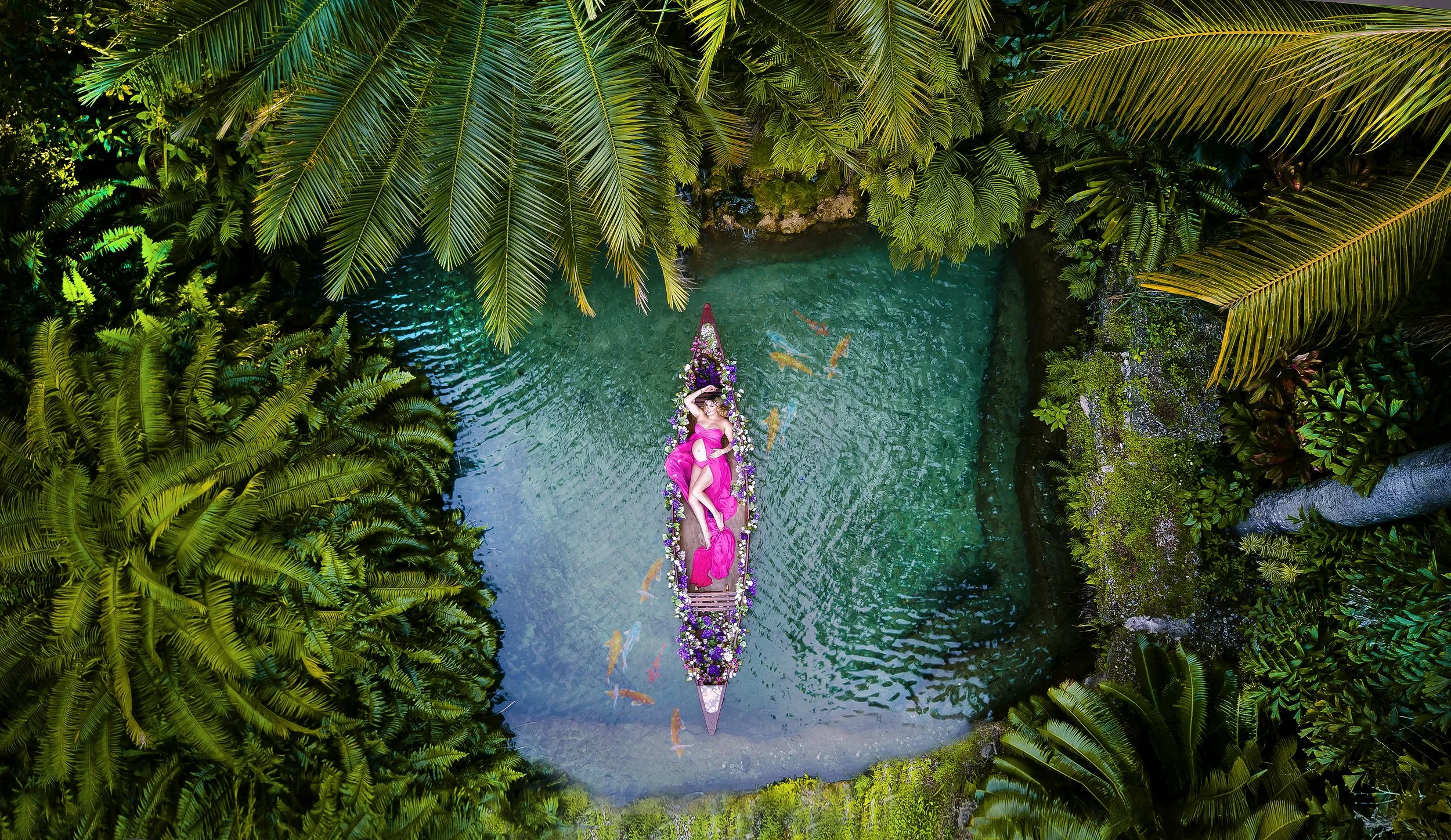 A woman in a pink dress lying on a decorated boat in a small turquoise pond surrounded by lush green tropical plants and trees.