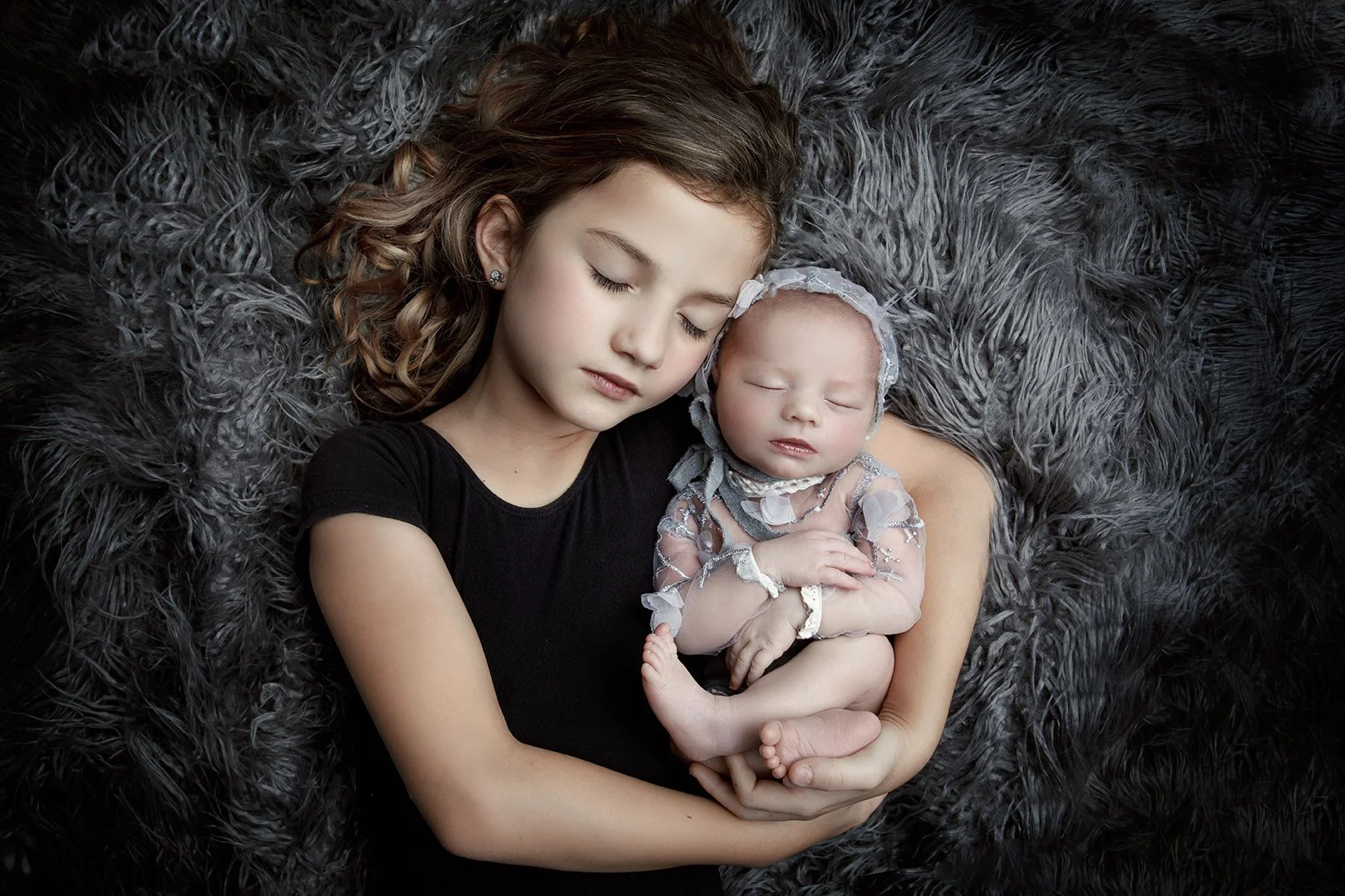 A young girl with light skin and curly hair is holding a baby with light skin, both with eyes closed, resting on a gray fuzzy blanket.