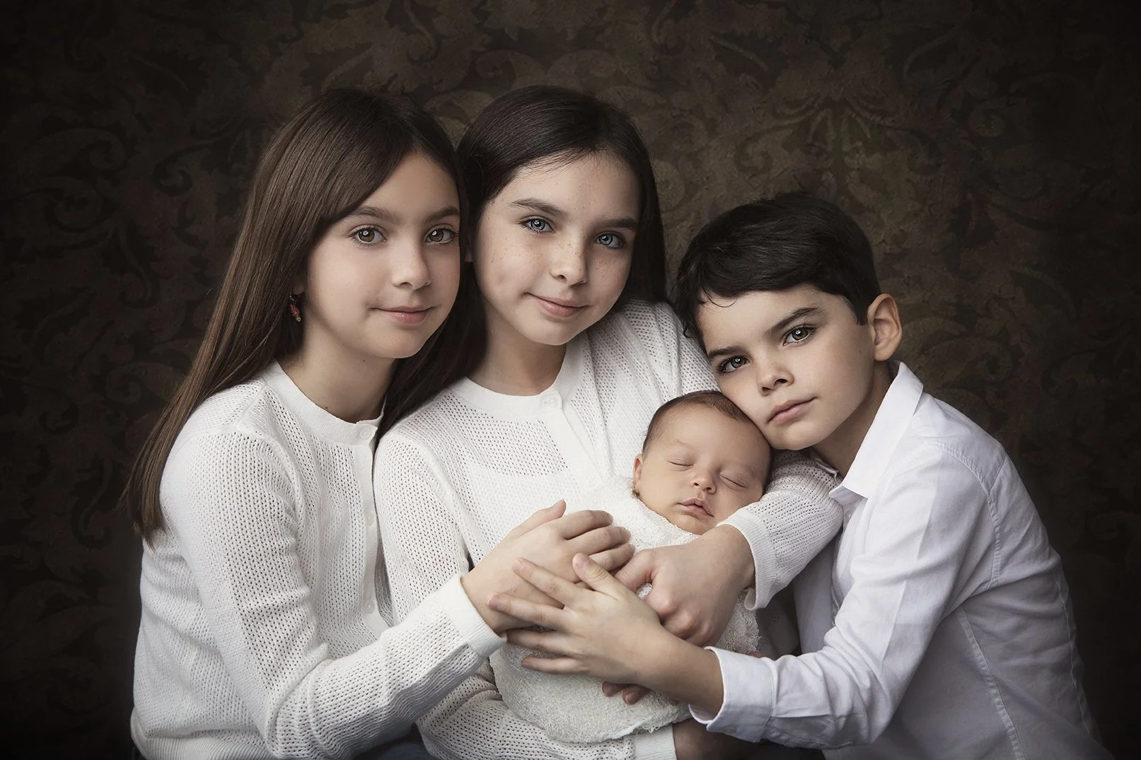 Four children, three girls and one boy, holding a sleeping baby, all dressed in white, posed against a dark patterned background.