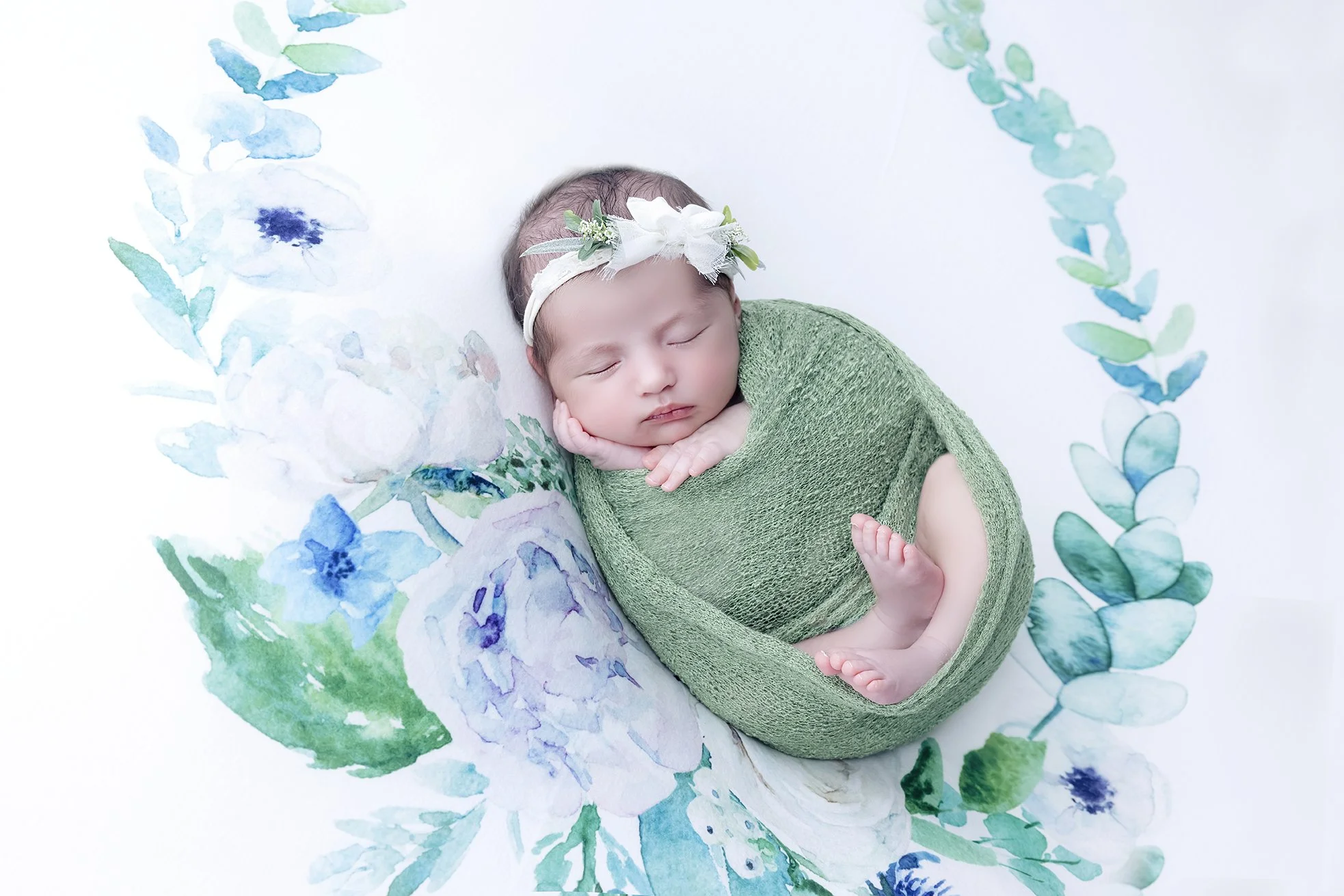 A sleeping baby wrapped in a green blanket with a white floral headband, lying on a white background decorated with watercolor blue and green flowers and leaves.