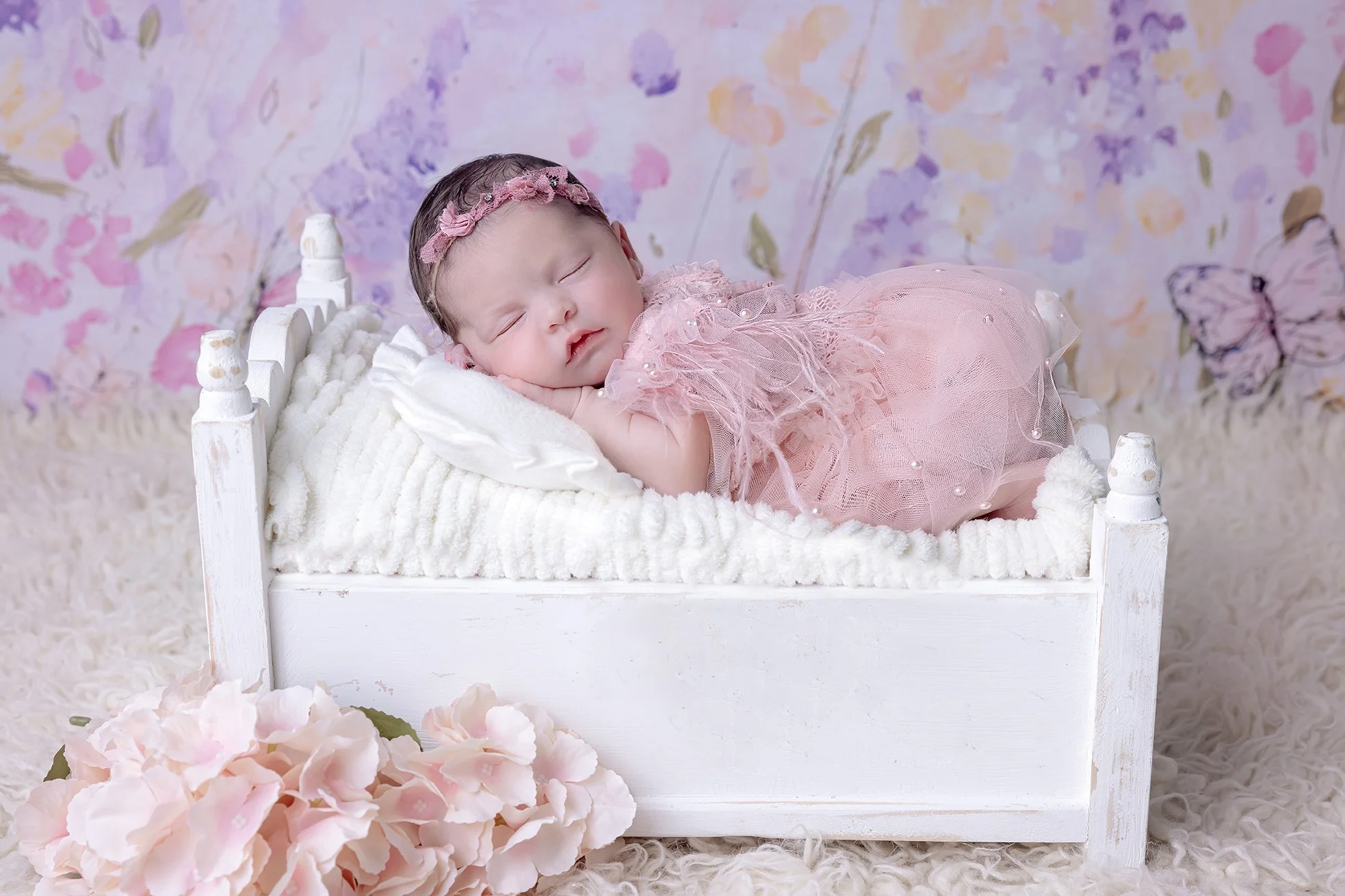 Sleeping baby girl in pink dress with floral headband on white bed with pink flowers and butterfly background.