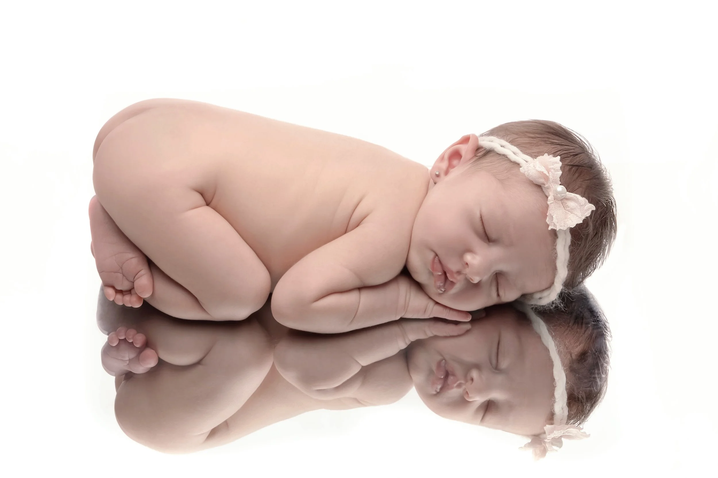 A sleeping newborn baby girl lying on her side with her hands under her cheek, wearing a pink lace headband with a bow and pearl accents, on a reflective white surface.