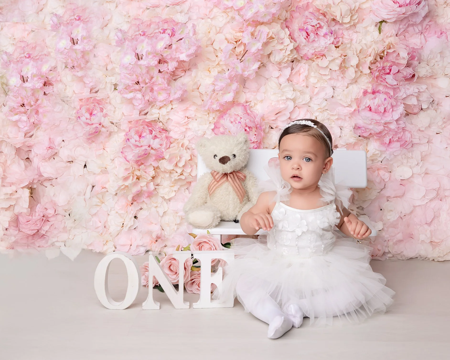 Young girl in a white dress sitting in front of a pink flower wall, with a teddy bear, a white chair, and white decorative letters spelling 'ONE' in front of her.