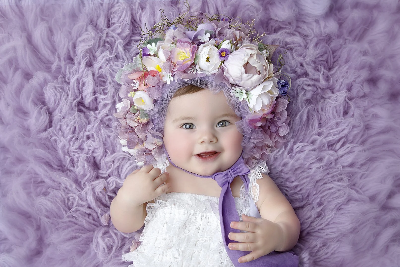 A smiling baby girl with blue eyes lying on a soft purple fur blanket, wearing a floral bonnet with pink, purple, and white flowers and a purple ribbon, dressed in a white lace dress.