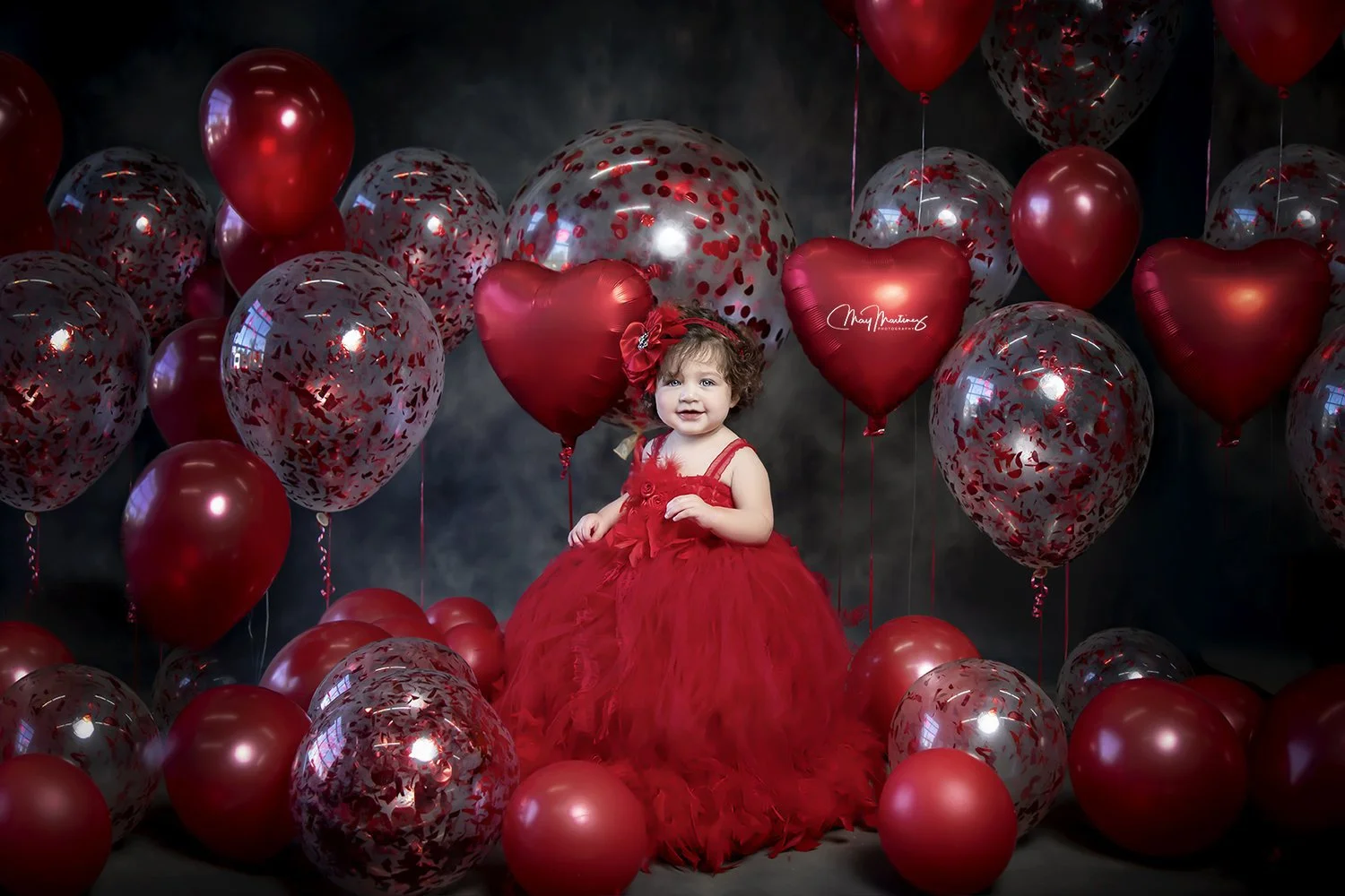 A young girl in a red dress and hair bow standing among red and confetti-patterned silver balloons against a dark background.