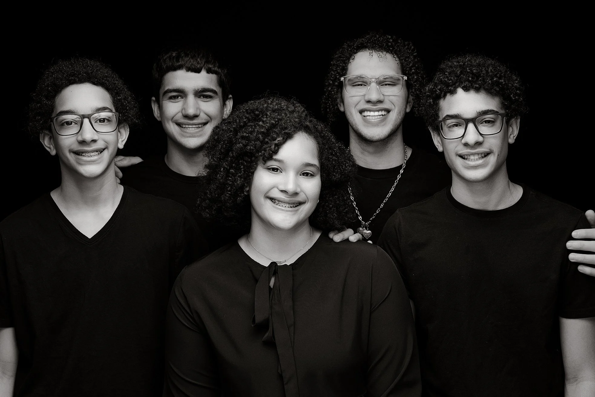 Group of six smiling teenagers with glasses and curly hair, posing against a black background.