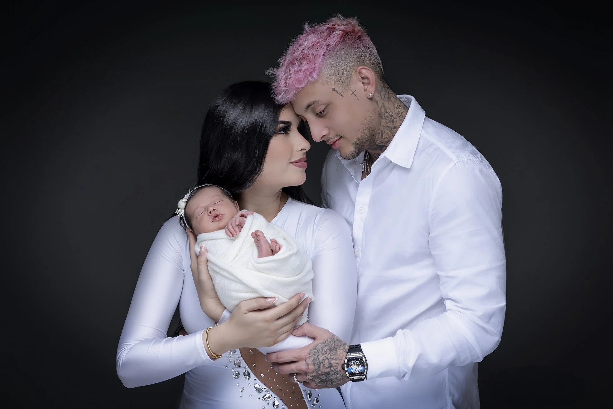 A family with a woman, a man, and a newborn baby in a studio with a dark background. The woman holds the baby, and the man is gently touching the woman’s stomach. The woman has long black hair and is wearing a white dress. The man has pink hair, tatt
