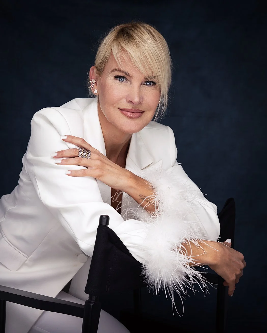 A blonde woman in a white blazer with feathered cuffs sitting on a black director's chair, looking at the camera with a slight smile.