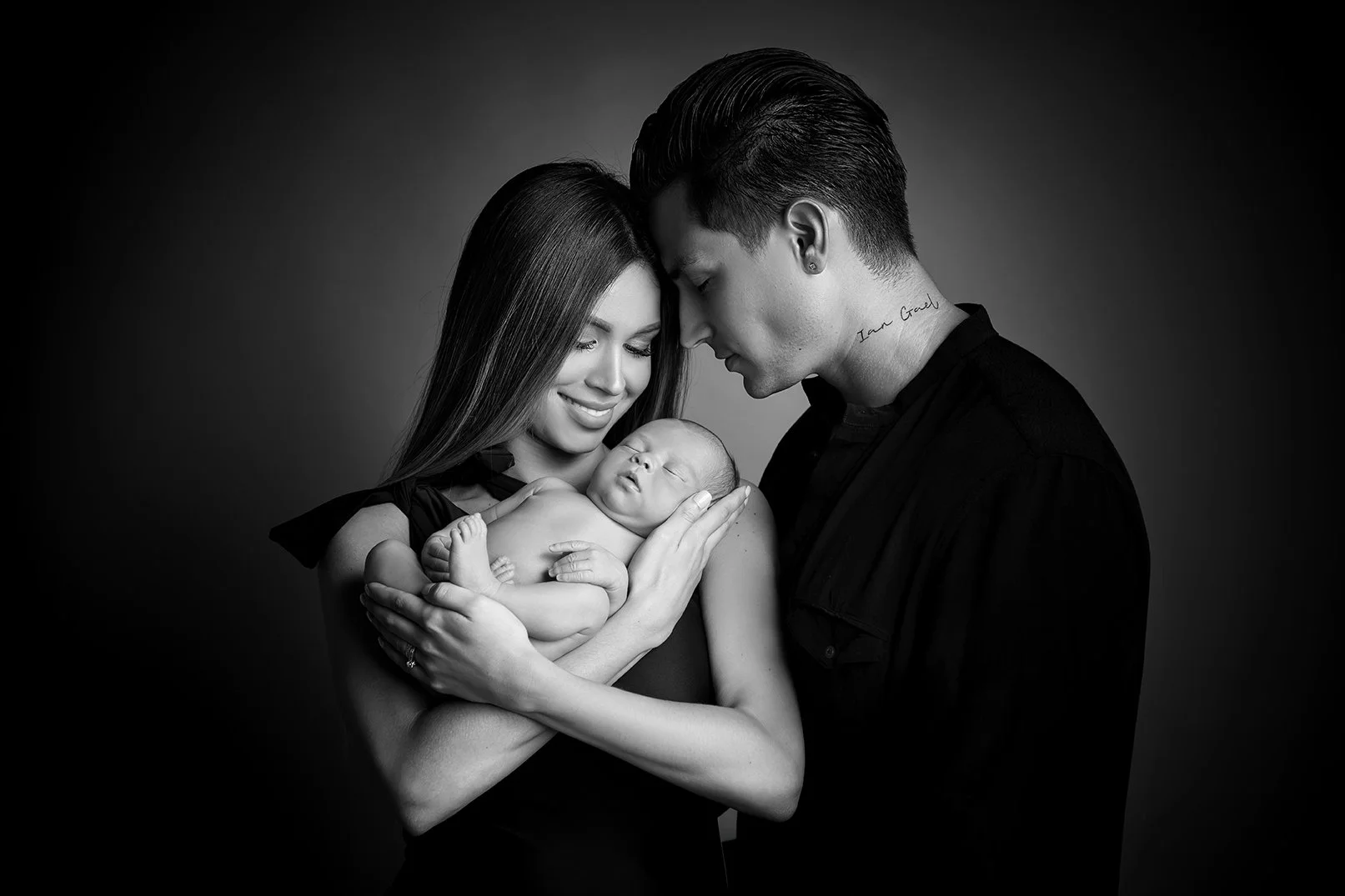 Black and white photo of a woman holding a newborn baby, with a man leaning in close, all with their foreheads touching, in a tender family portrait.