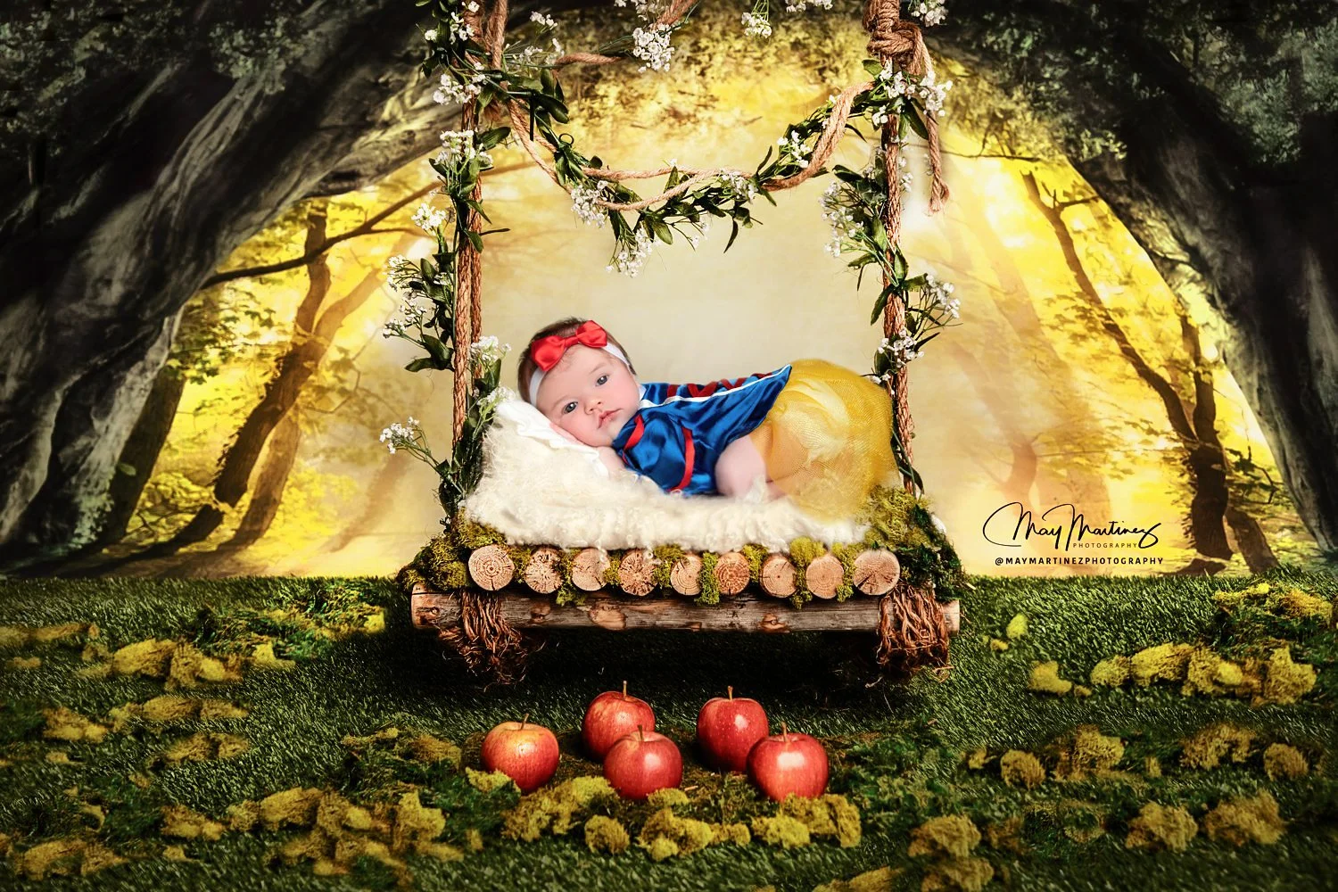 Baby dressed in a Snow White costume lying on a swing in a woodland setting with apples on the ground in front.