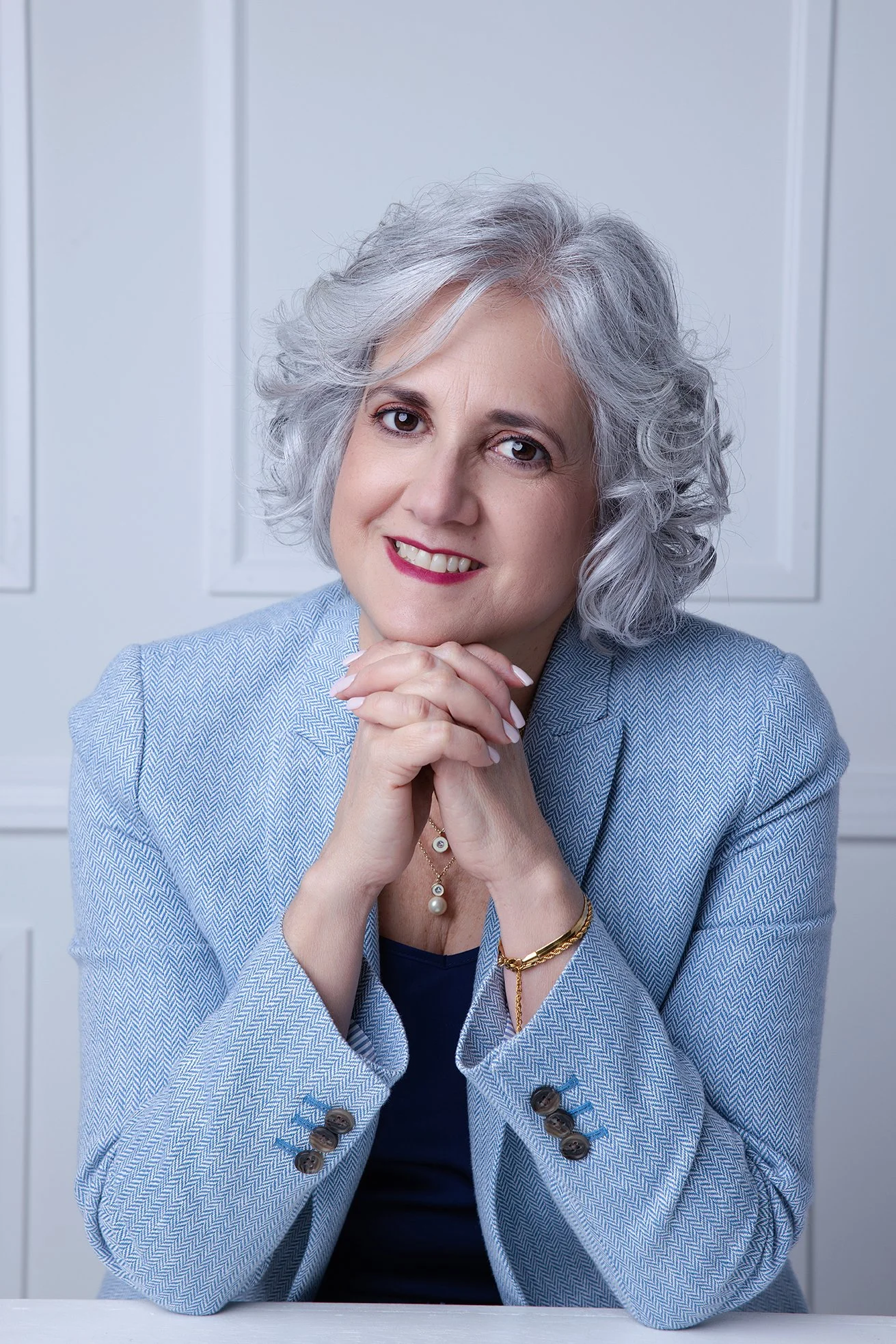 Portrait of a smiling middle-aged woman with gray curly hair, wearing a light blue blazer and sitting with her hands clasped under her chin against a white paneled background.