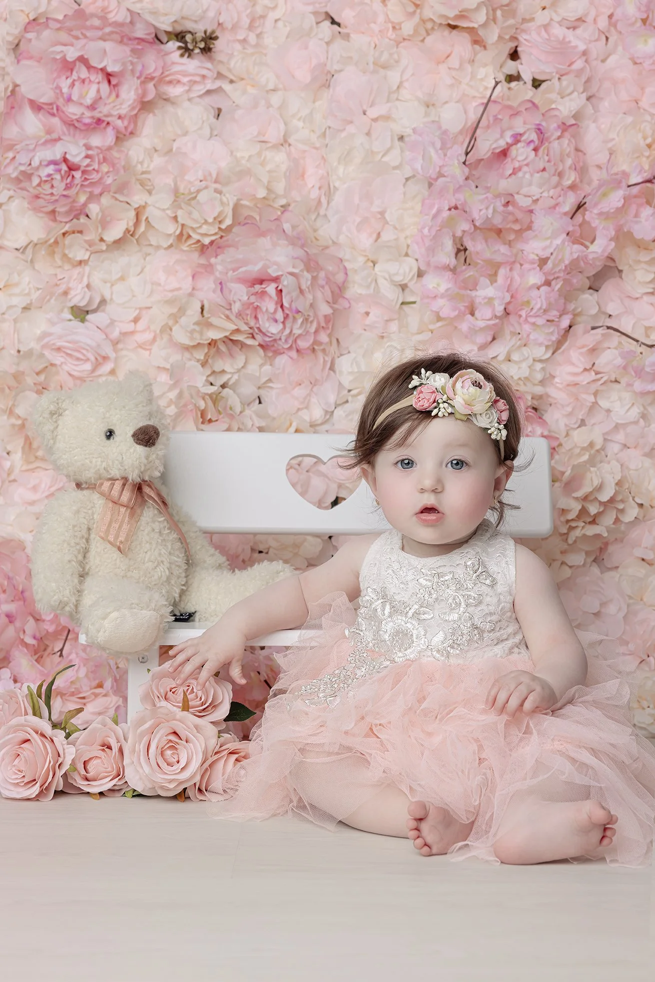 A young girl wearing a floral headband and a pink tulle dress sitting on the floor in front of a wall decorated with pink flowers, with a teddy bear and pink roses nearby.