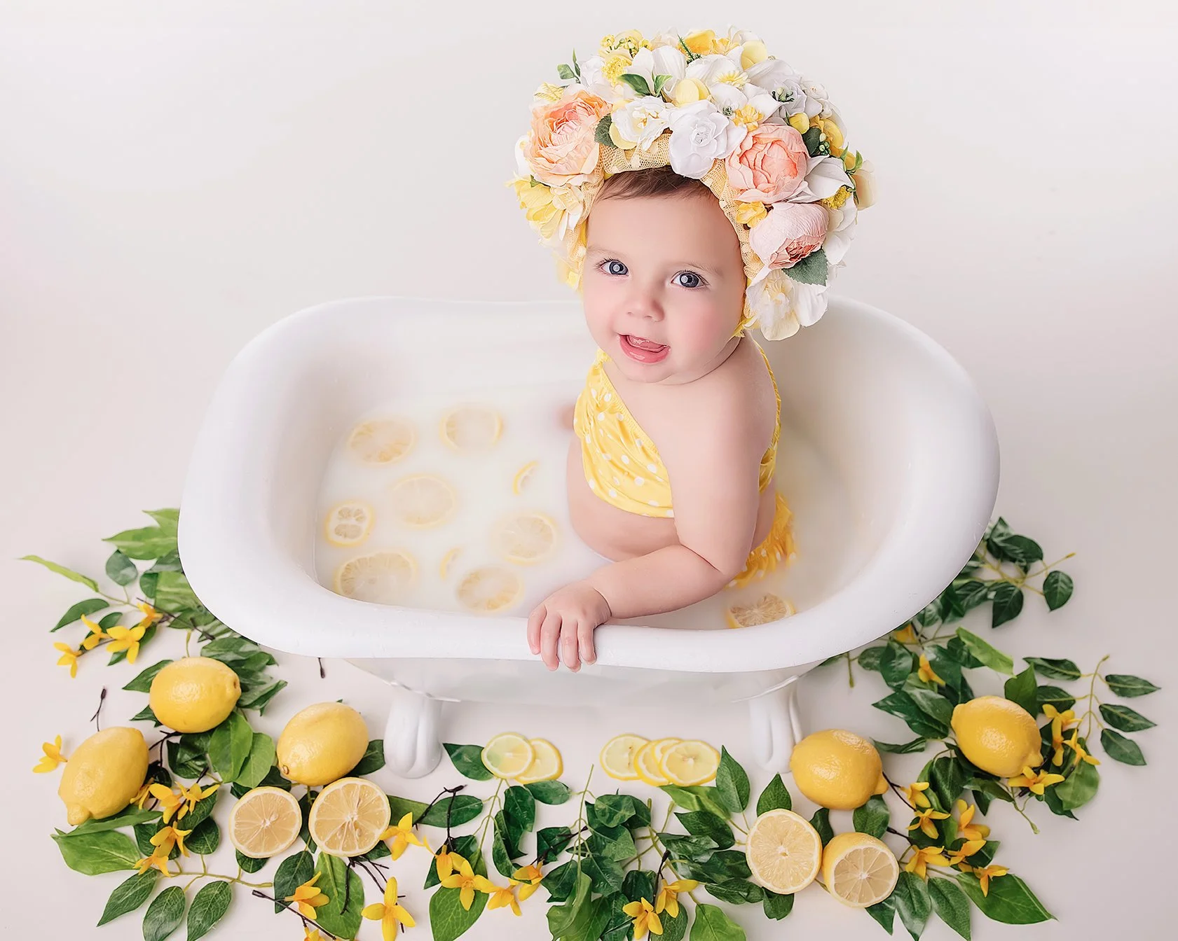 A smiling baby girl with blue eyes sitting in a small white bathtub filled with lemon slices and milk, wearing a yellow polka dot swimsuit and a large flower wreath on her head, surrounded by lemons and yellow flowers.