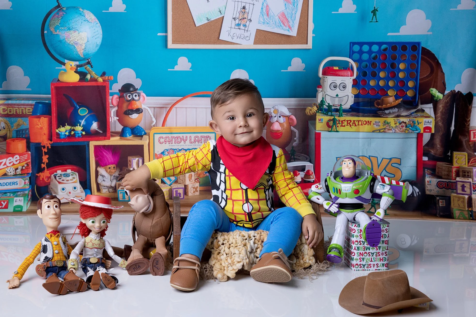 Young boy dressed as Woody from Toy Story, sitting among Toy Story toys including Buzz Lightyear and Jessie, with various other toys and colorful backdrop featuring clouds, a globe, and drawings on the wall.