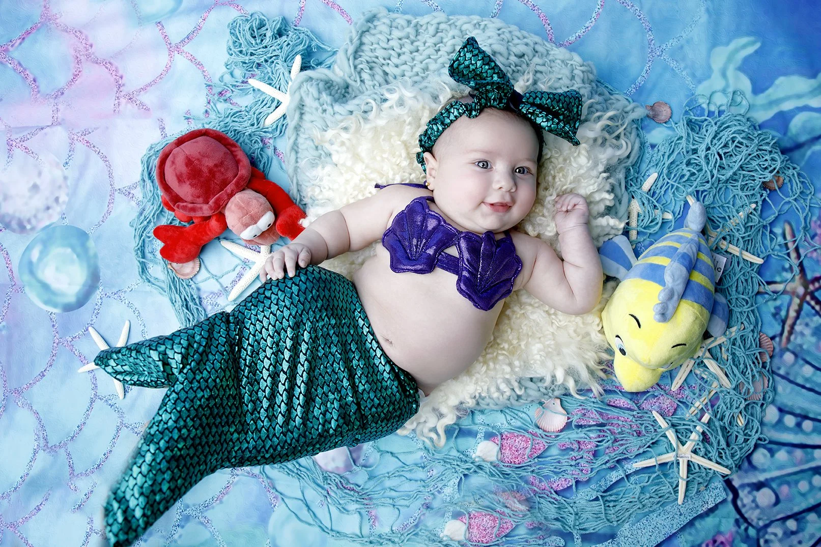 Baby dressed as a mermaid lying on a blue and white ocean-themed blanket, surrounded by plush sea creatures like a red crab, a yellow and blue fish, shells, and starfish.