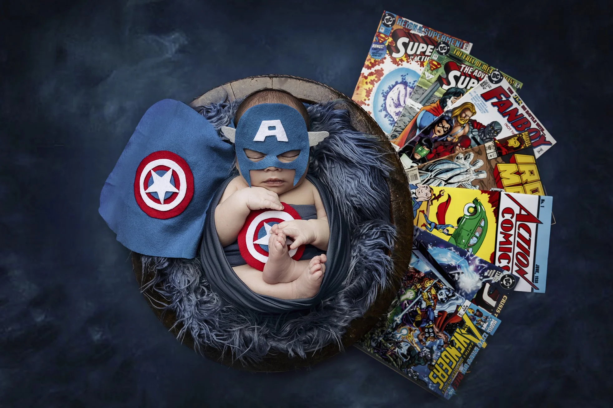 A baby dressed as Captain America, wearing a blue mask with an 'A' and wings, lying on a fluffy gray blanket. The baby holds a small Captain America shield in their hand. Surrounding the baby are various comic books with superhero themes, spread out 