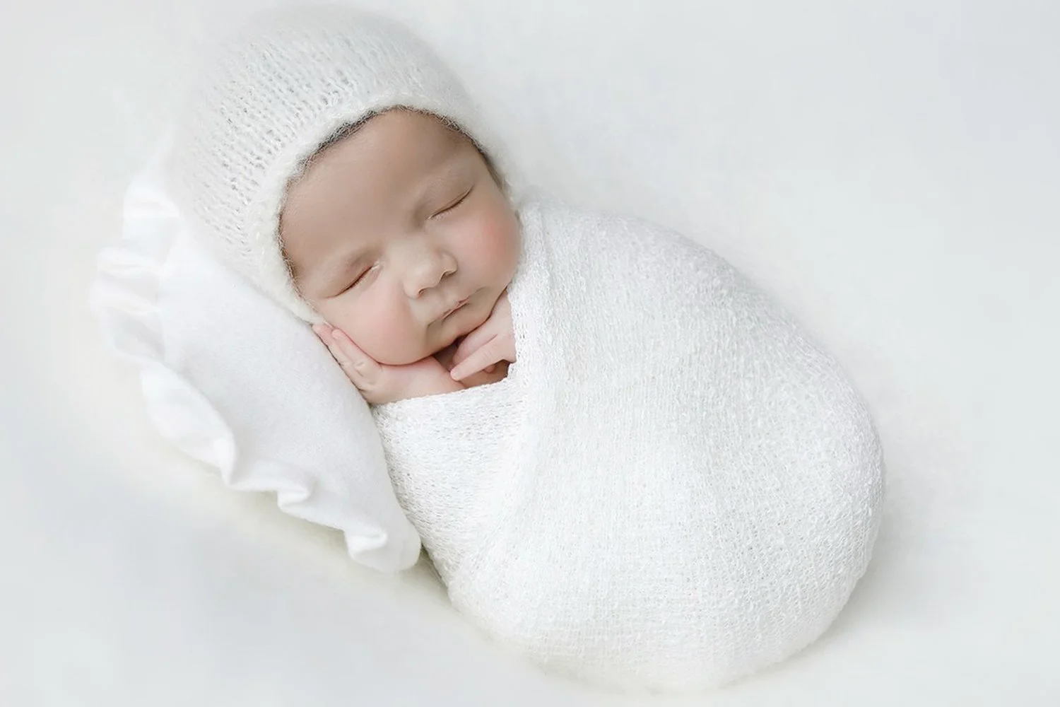 A sleeping newborn baby wrapped in white cloth, wearing a knitted white hat, resting on a white pillow.