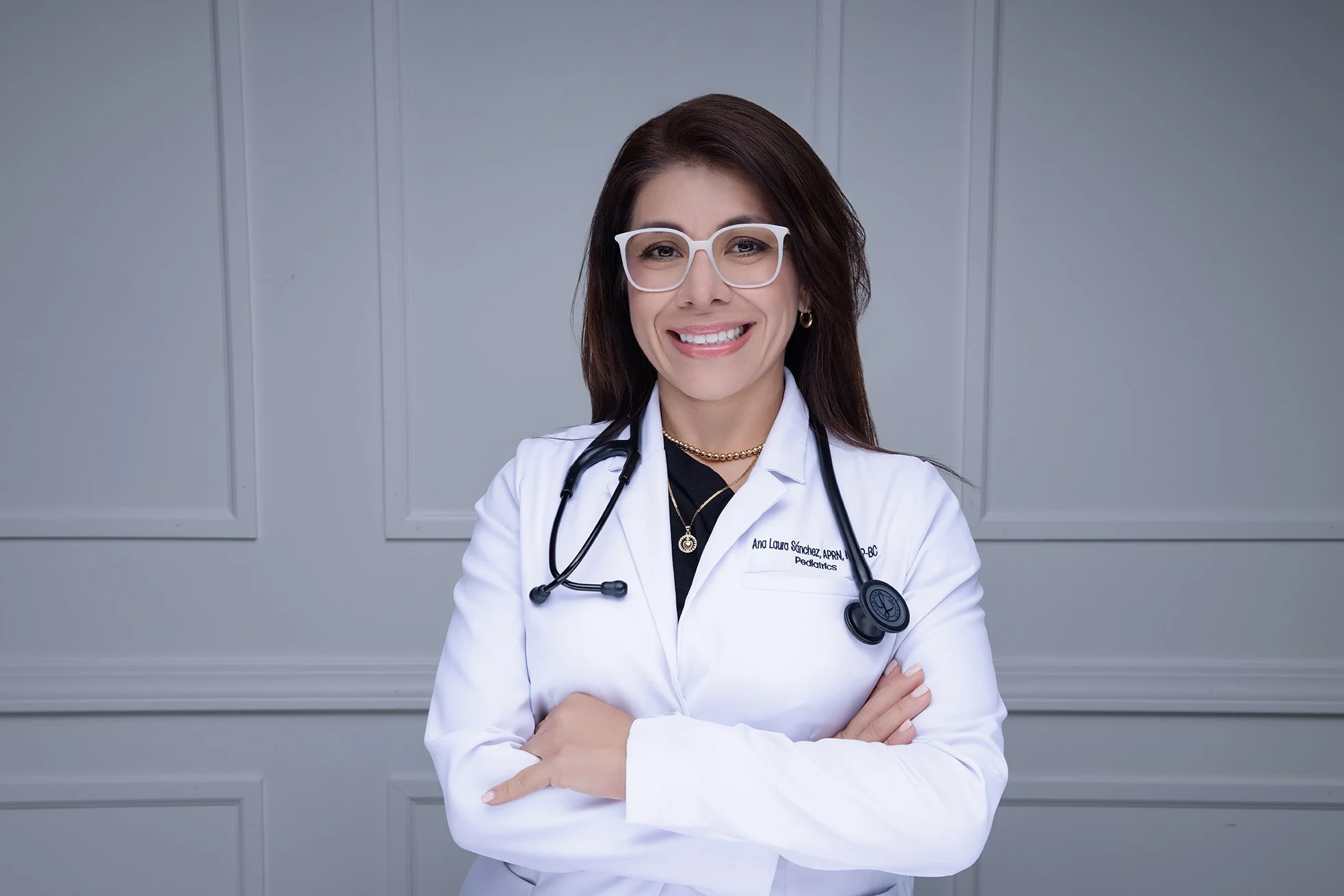Female doctor wearing glasses and a white coat with a stethoscope around her neck, standing with arms crossed and smiling in a professional setting.