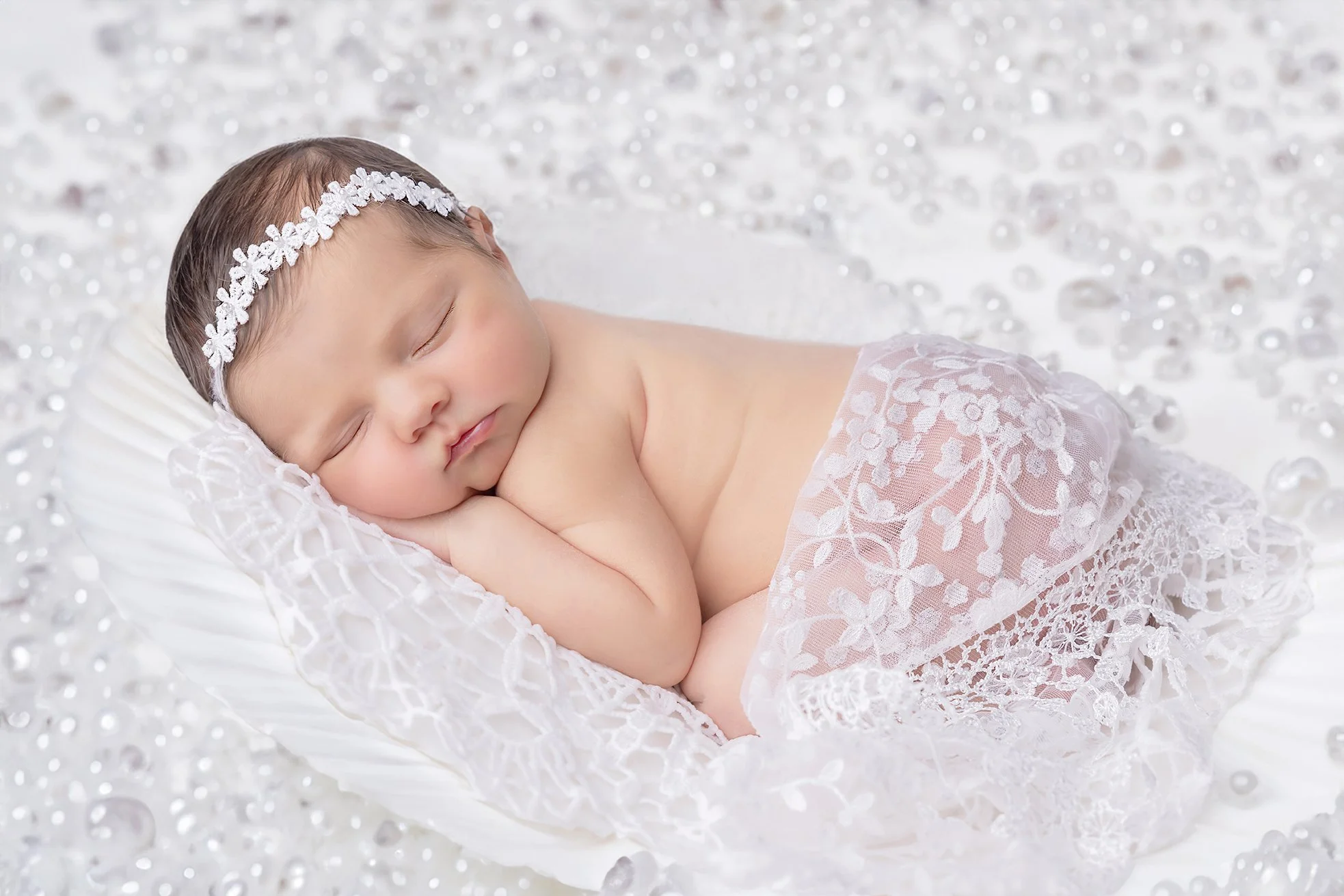 A sleeping baby girl with a headband, wrapped in lace, on a soft cushion surrounded by silver beads.