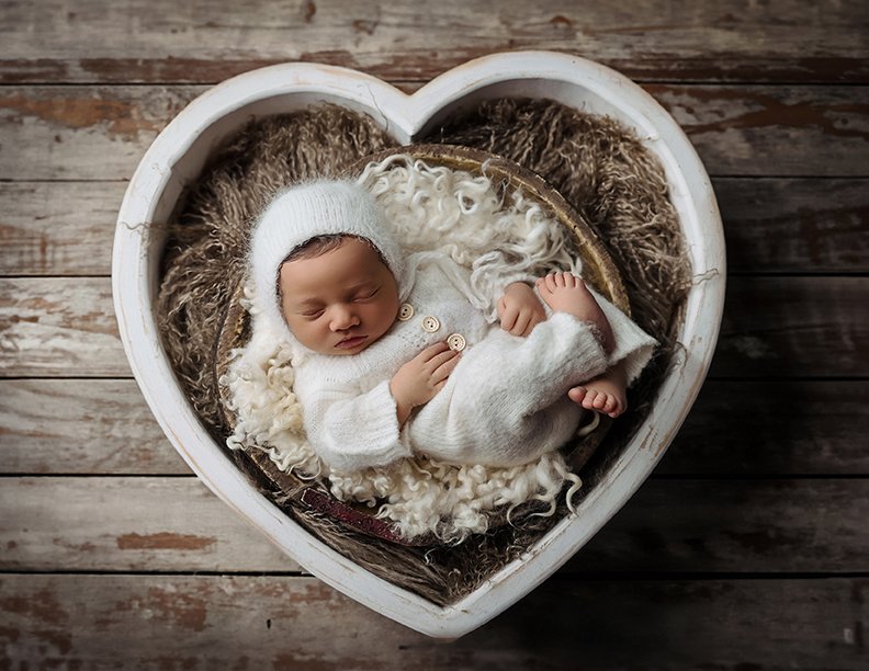 A newborn baby peacefully sleeping in a heart-shaped white box, surrounded by soft wool blankets and plush fabric, on a wooden floor.