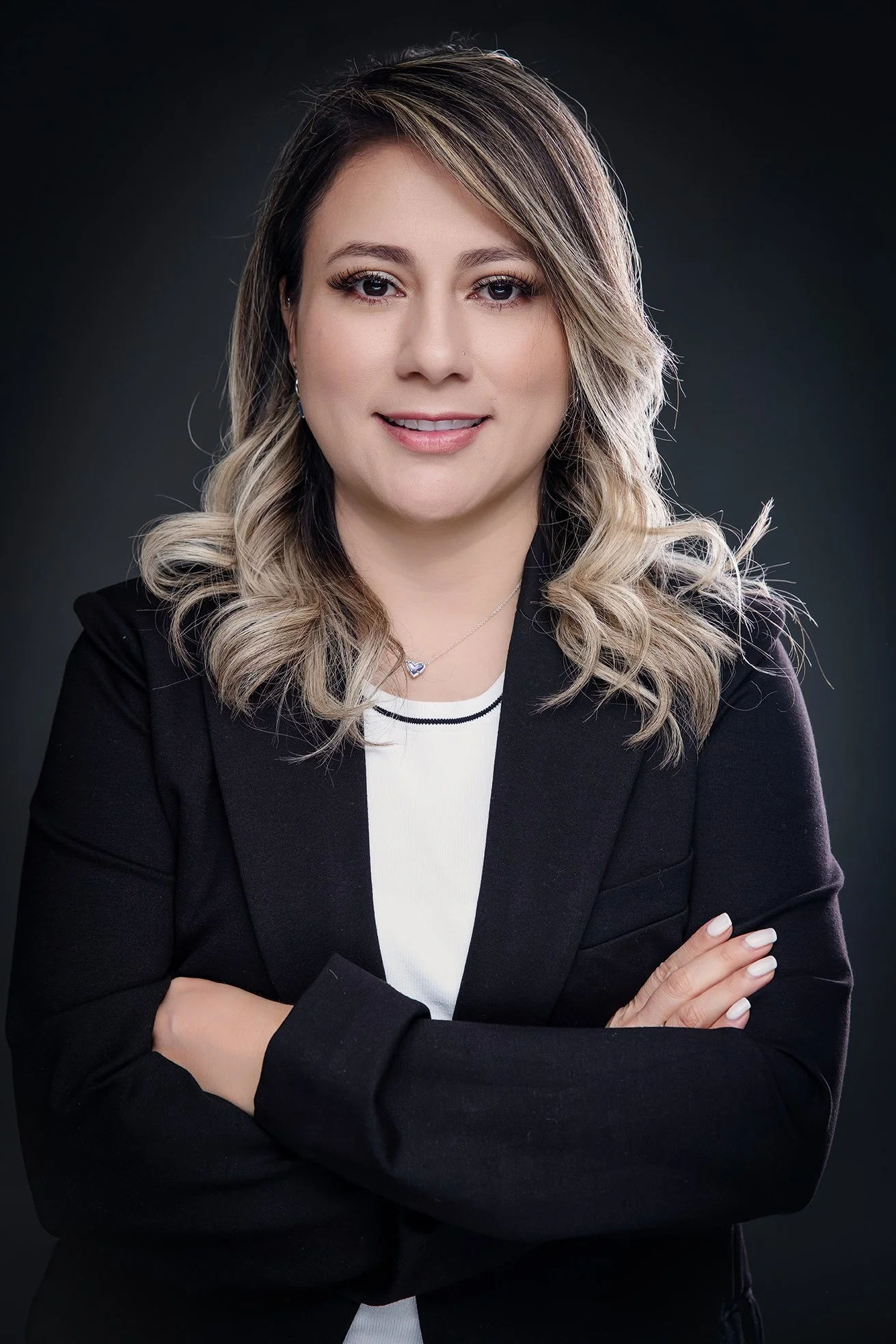 A woman with blonde wavy hair, wearing a black blazer and white top, smiling with arms crossed against a dark background.