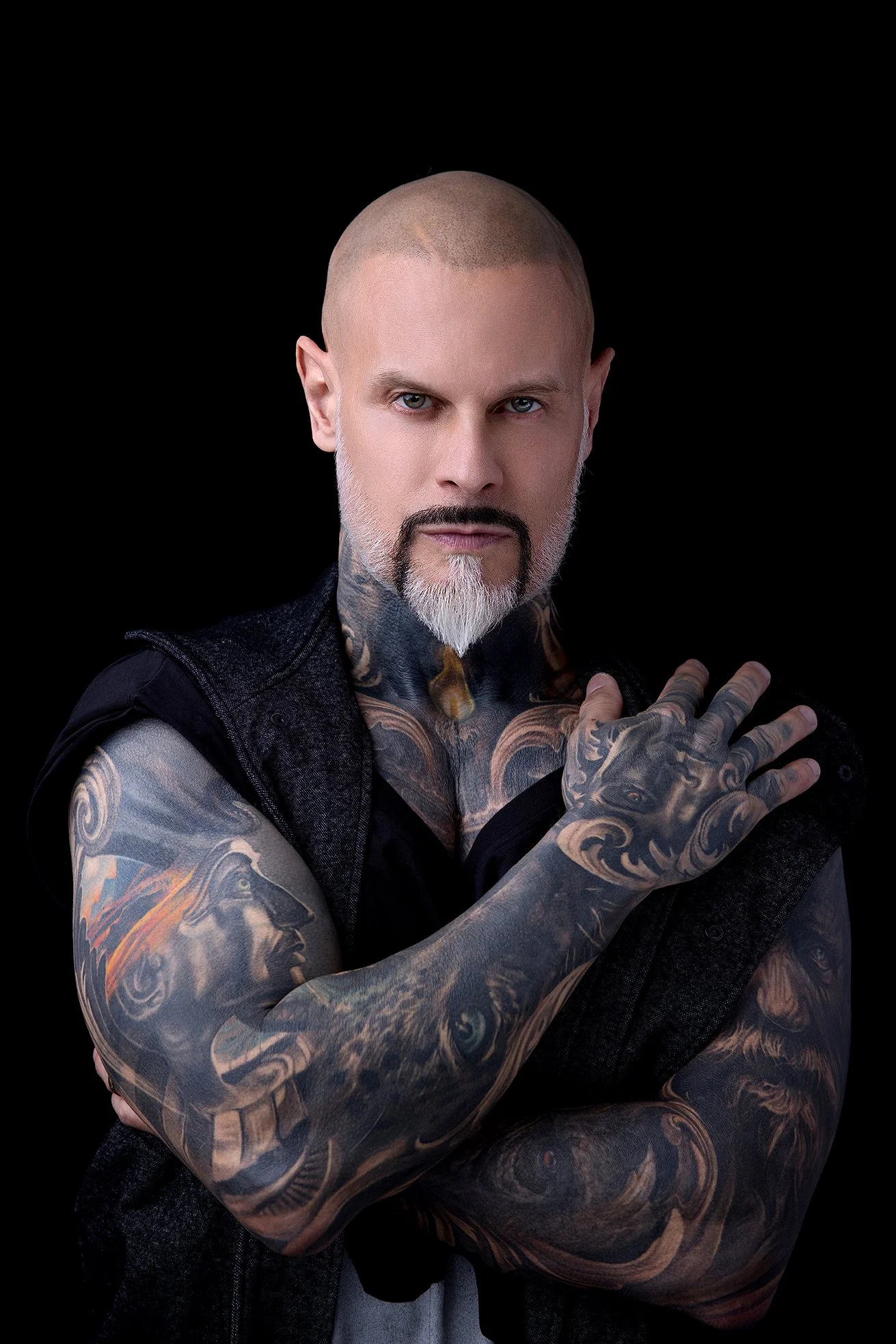A tattooed man with a shaved head, white beard and intense expression posing against a black background.