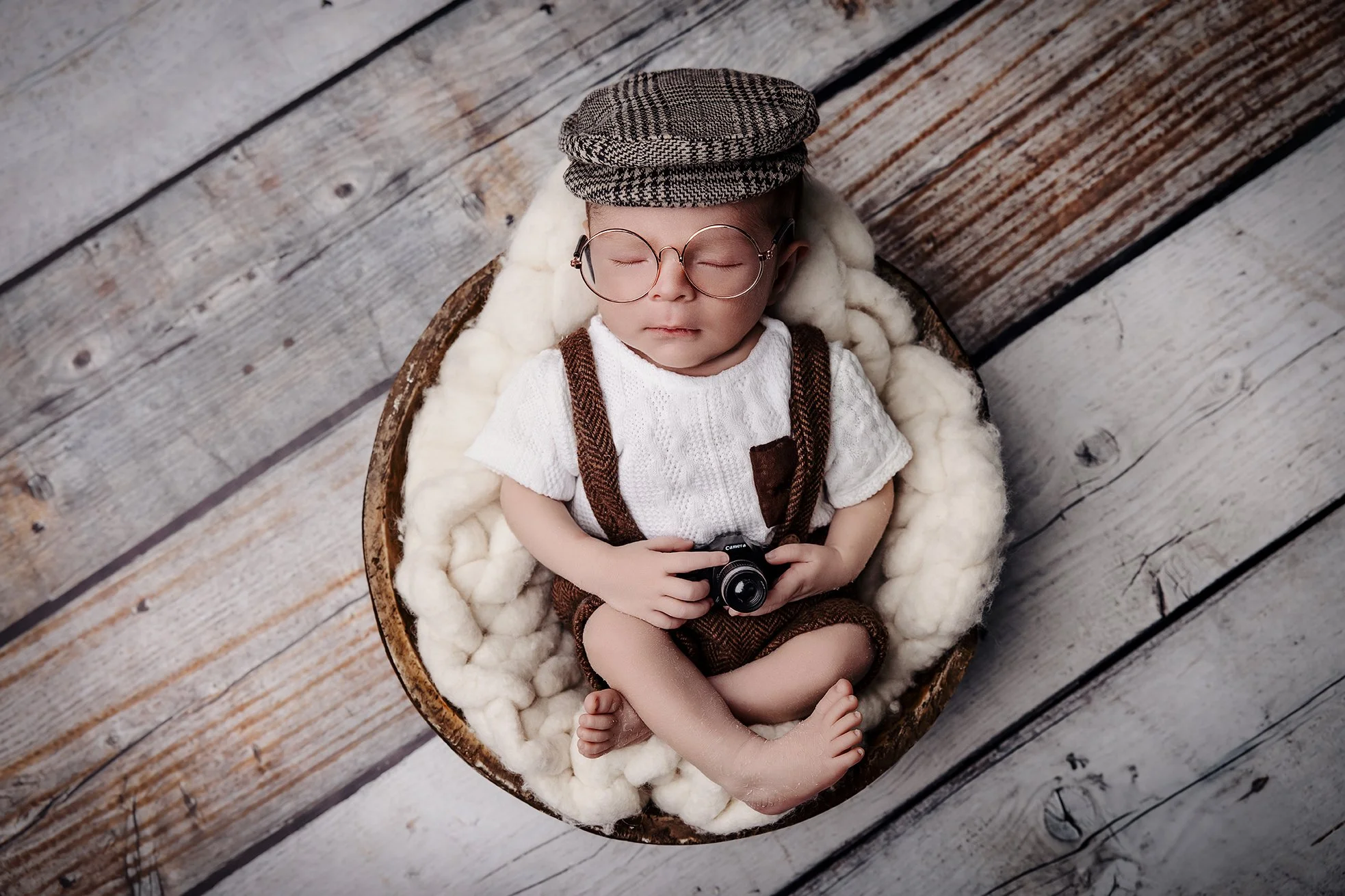 A young child with glasses, a flat cap, and vintage-style clothing, sitting in a wooden bowl lined with soft white blanket, holding a small camera, on a wooden floor.