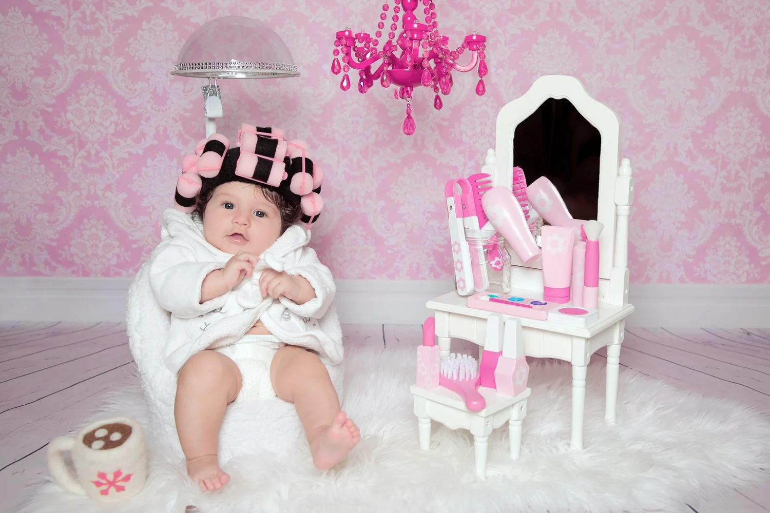 Baby sitting on a white fluffy rug in a pink-themed room with a pink chandelier, a mirror, and a toy vanity with pink and white accessories.