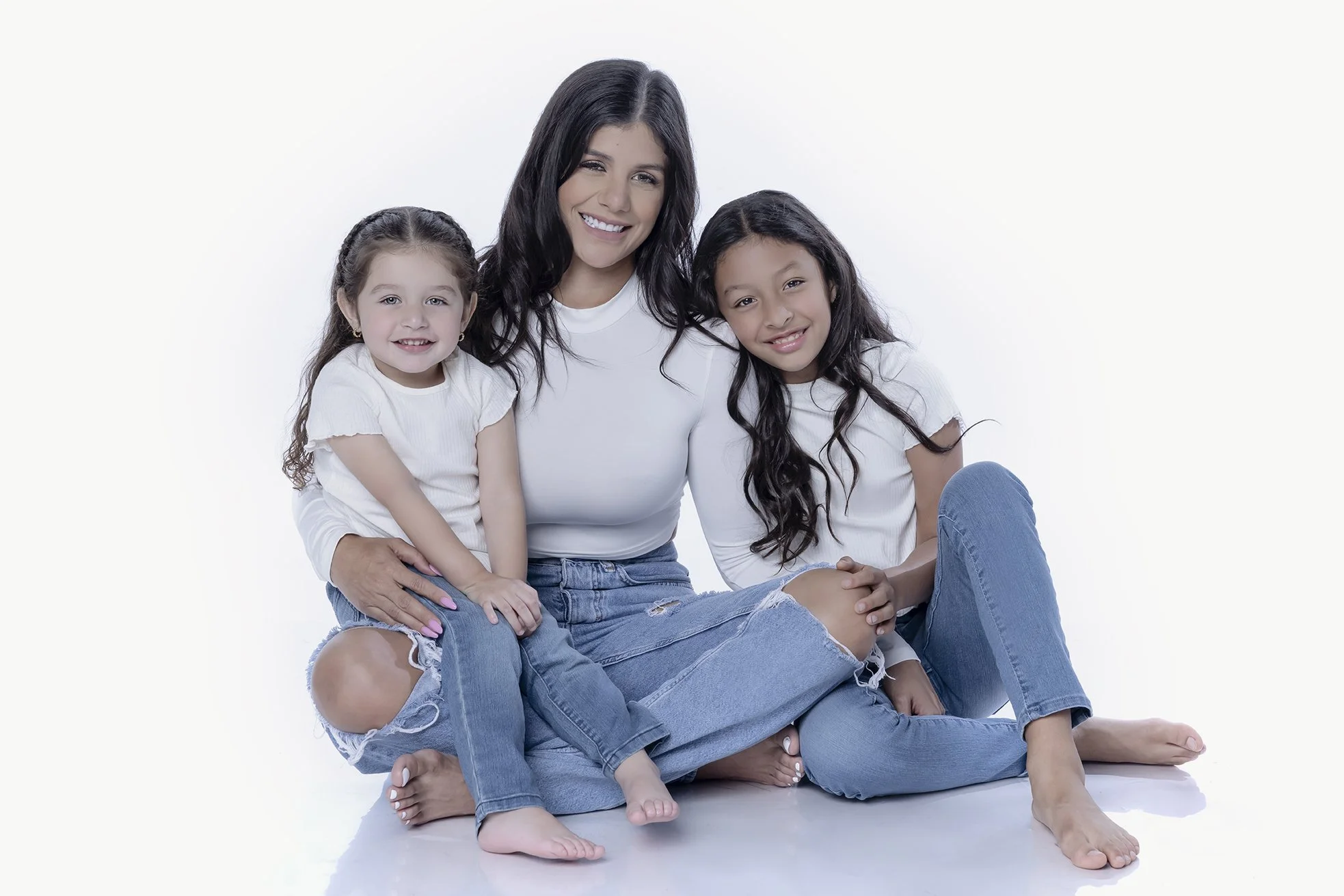 A woman and two young girls sitting on the floor in white shirts and blue jeans, smiling and posing for the photo against a plain white background.