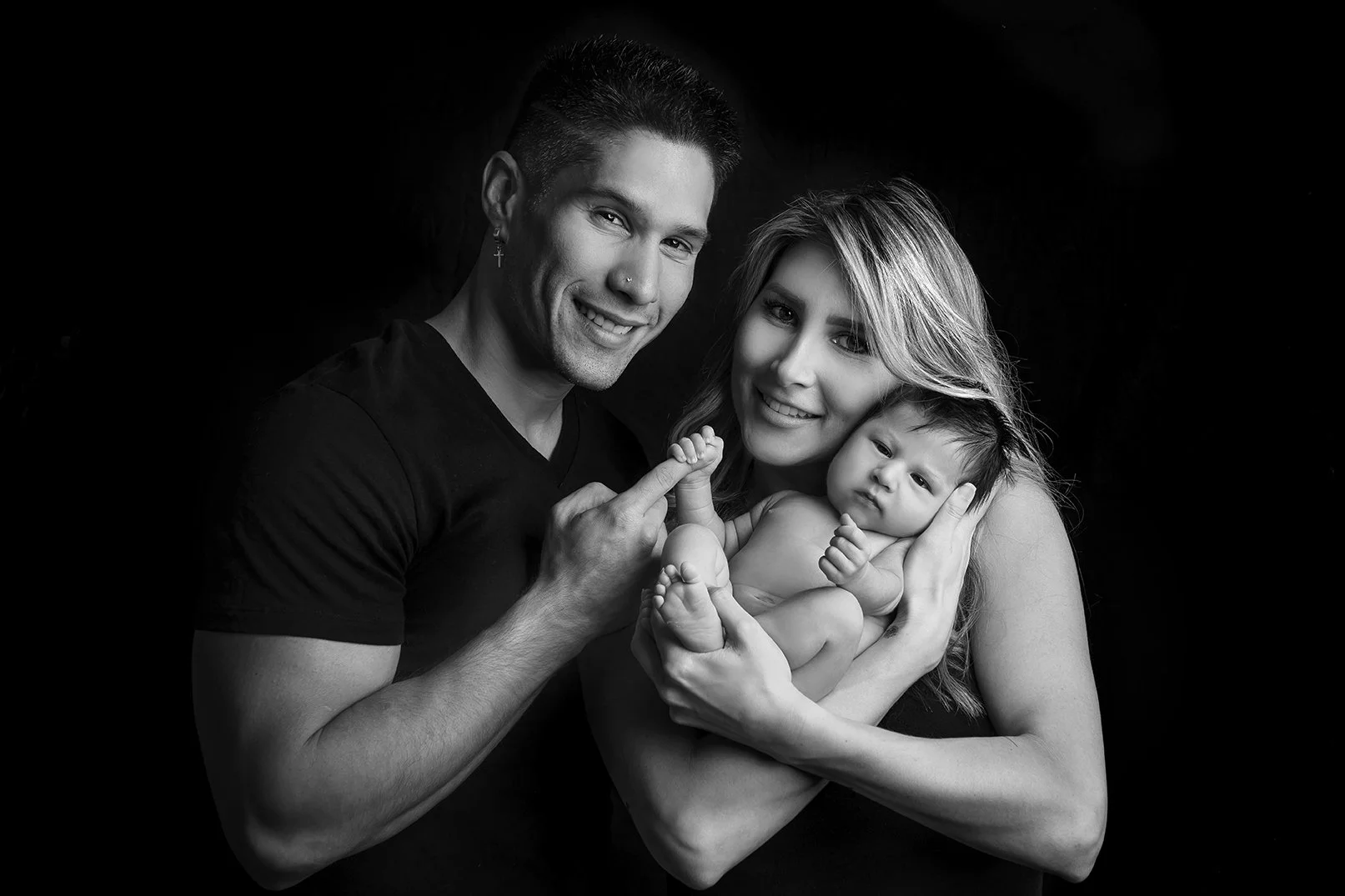 A black-and-white portrait of a happy family of three with a man, a woman, and a baby. The man is on the left, smiling and holding up his middle finger. The woman is in the center, gently holding the baby, who is resting in her arms and looking at th