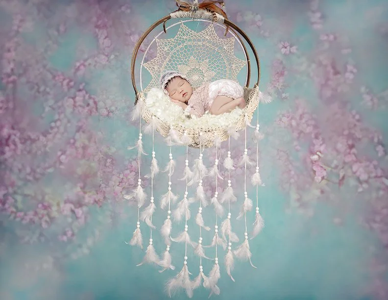 A newborn baby sleeping in a hanging dreamcatcher with a floral patterned backdrop.