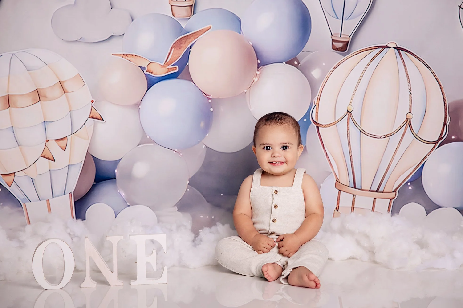 Young child sitting on the floor in front of a backdrop with hot air balloon and cloud illustrations, surrounded by balloons and cotton resembling clouds, with white decorative letters spelling 'ONE' indicating a first birthday celebration.