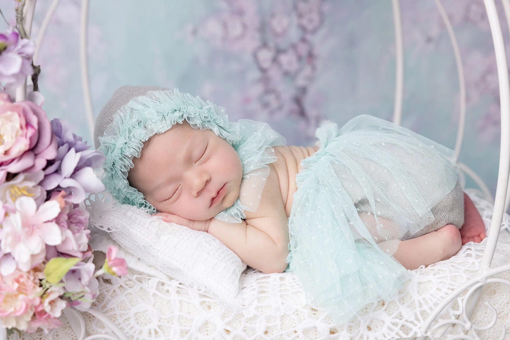 A sleeping baby lying on a white lace blanket in a small white bed, with pink and purple flowers on the side and a pastel-colored background with cherry blossom trees