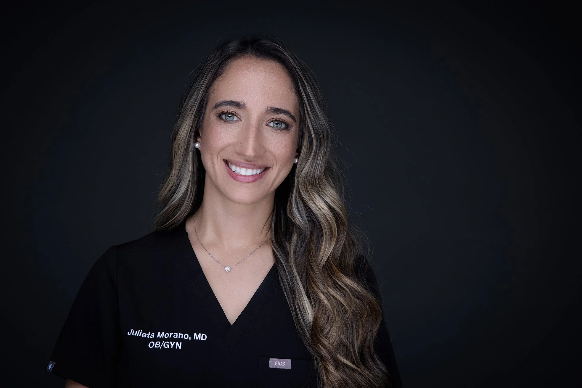 Professional portrait of Dr. Julieta Morano, a woman with long wavy hair, wearing a black medical uniform with her name and credentials embroidered on it, and smiling against a dark background.