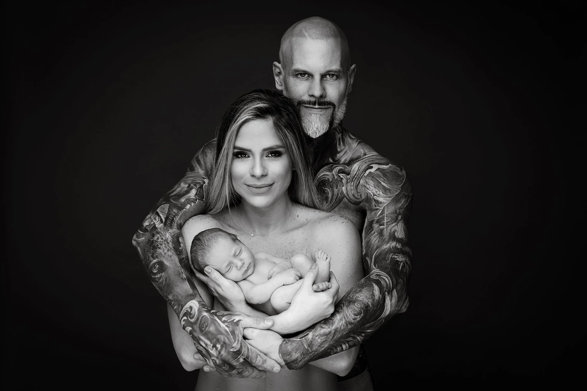 A black and white photo of a man with tattoos holding a woman and a sleeping baby. The man is standing behind the woman with his arms around her, and they are all looking at the camera.