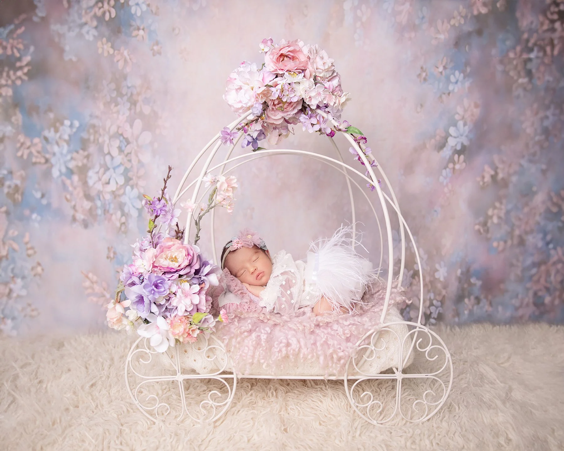 A sleeping baby girl lying on a pink blanket inside a white, decorative, carriage-shaped bed adorned with pink and purple flowers. The background is a pastel floral pattern.