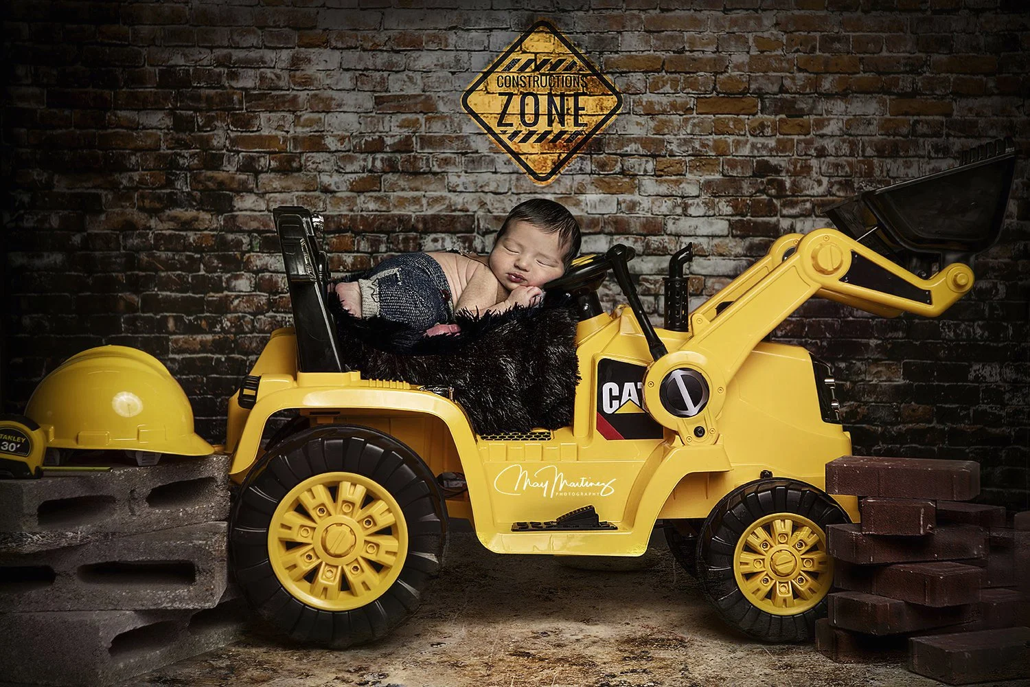 A sleeping baby lying on top of a yellow toy construction vehicle, with a black furry blanket underneath them. The background features a brick wall with a yellow and black construction zone sign. There are bricks and cinder blocks on either side of t