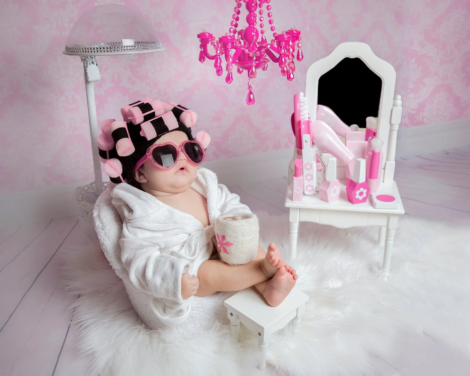 Baby dressed in a robe with curlers in her hair and wearing pink sunglasses, sitting on a fluffy rug next to a toy vanity table, with pink chandelier and pink wallpaper in the background.