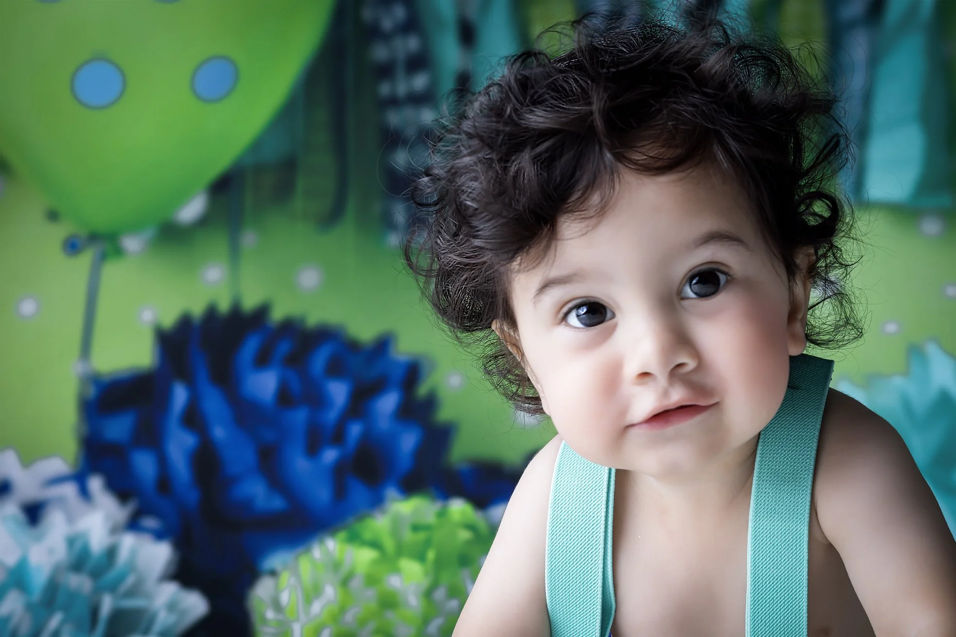 A young child with curly dark hair and big eyes looking at the camera, with colorful balloons and decorations in the background.