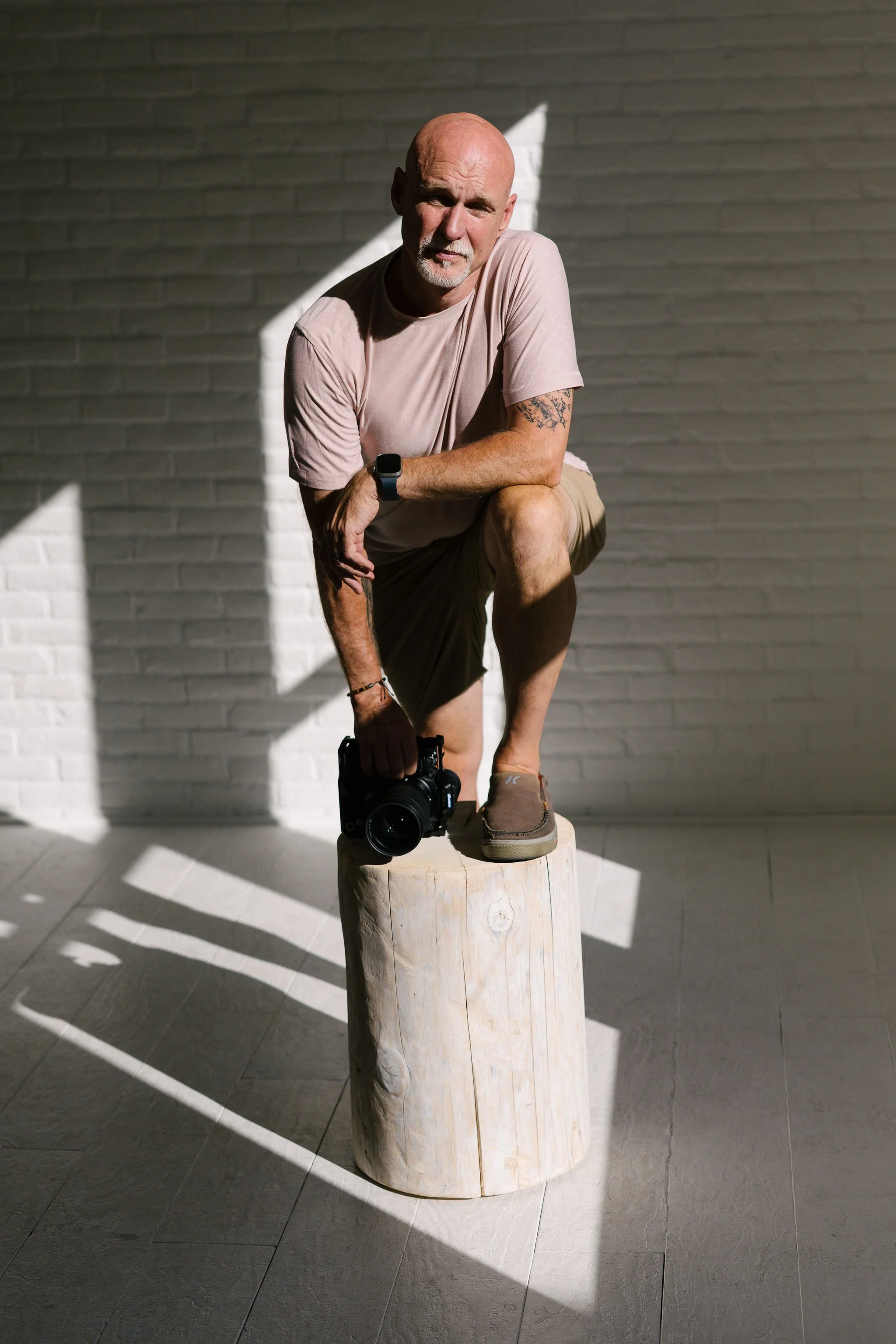 A bald, middle-aged man wearing a light pink t-shirt and beige shorts is crouching on one knee on a wood stump indoors. He has a tattoo on his left arm and is holding a camera in his right hand. He is looking at the camera with a serious expression. Sunlight is casting shadows on a white brick wall behind him.