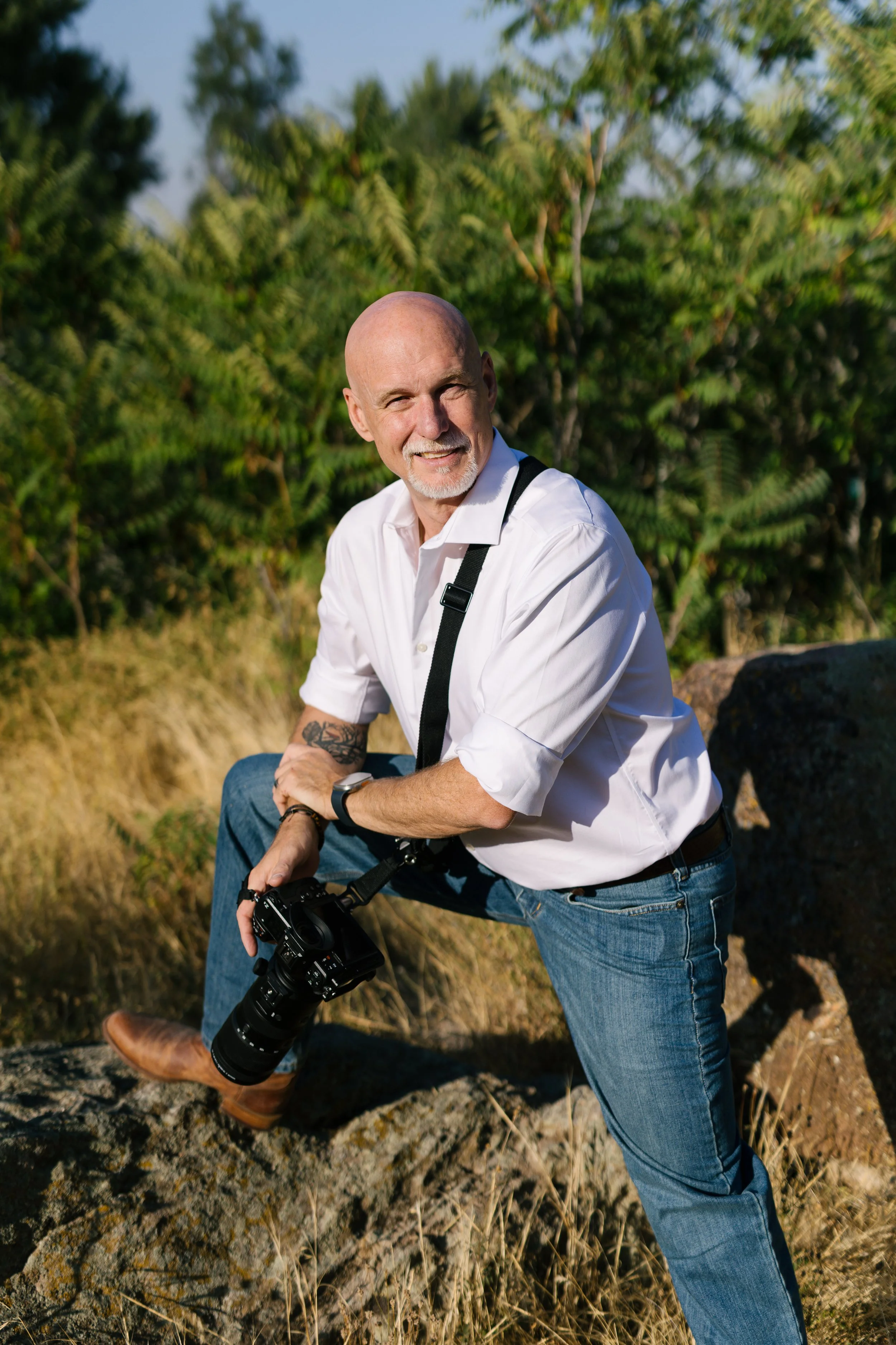 Bald man with a goatee smiling outdoors, holding a camera, dressed in a white shirt and jeans, sitting on a log in a natural setting with trees and grass.