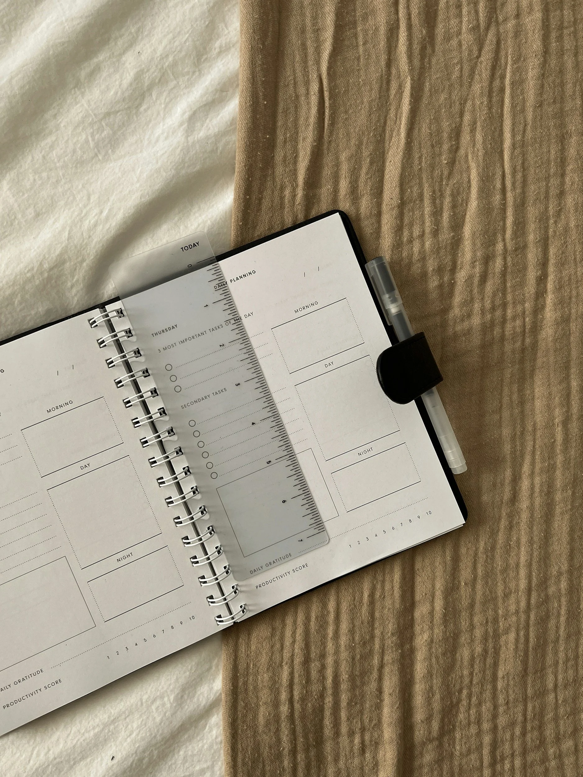 Open planner with daily task sections, a clear ruler, and a pen, laid on a wooden surface with a beige cloth nearby.