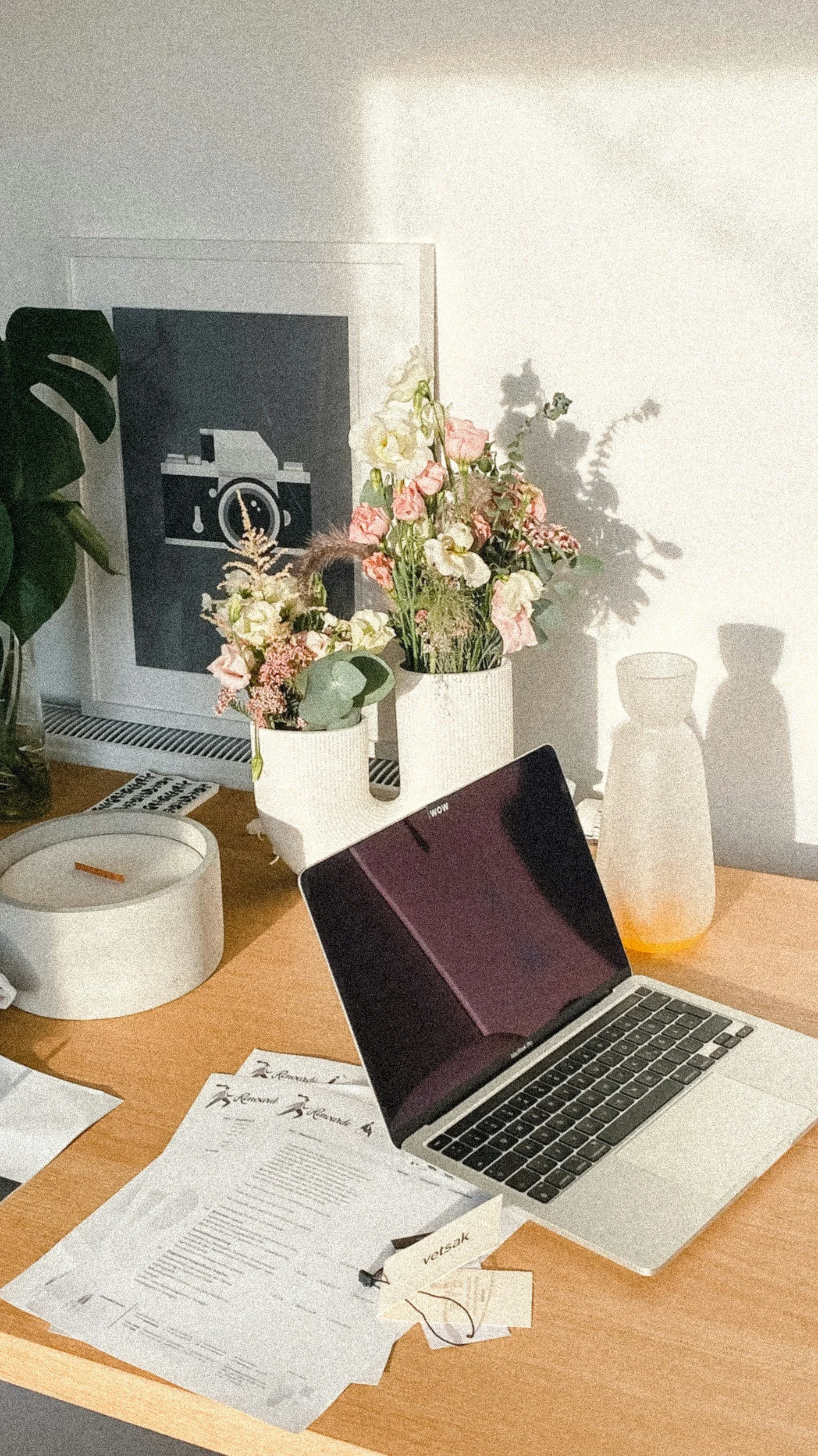 Desk with laptop, papers, a white candle, two vases with pink and white flowers, a framed camera artwork, and a large plant to the left. Shadows cast on the wall behind.