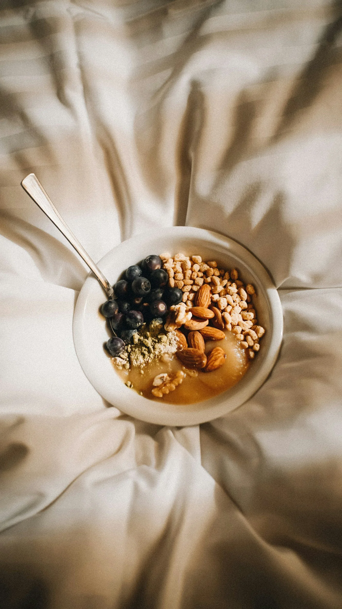 Top-down view of a white bowl with blueberries, almonds, granola, and honey on a cream-colored fabric background.