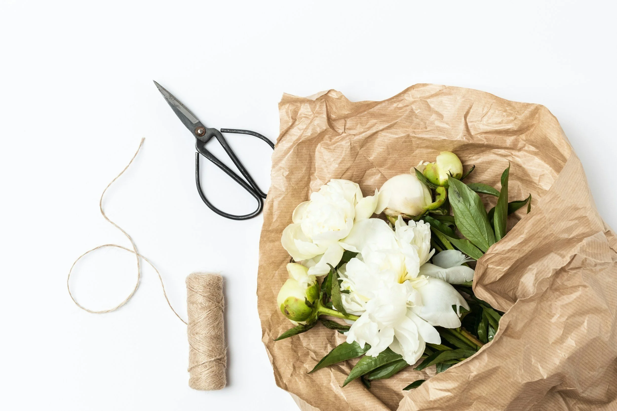 White peonies on brown paper with gardenias, scissors, twine, and a spool of string on a white background.