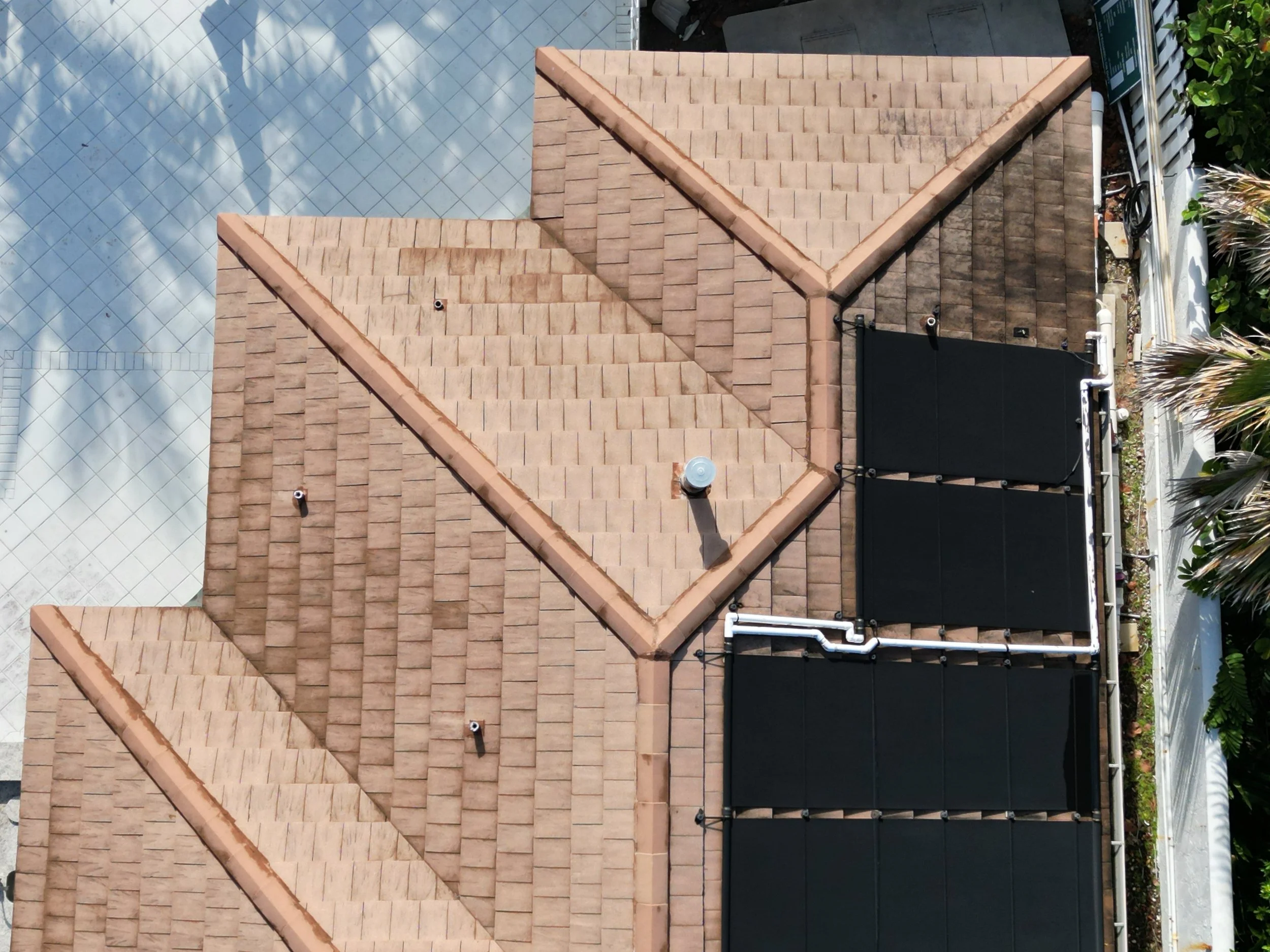 An aerial view of a house roof with brown shingles, some vents, and a black solar panel. On the right side, there is a white fence with green plants and trees nearby.