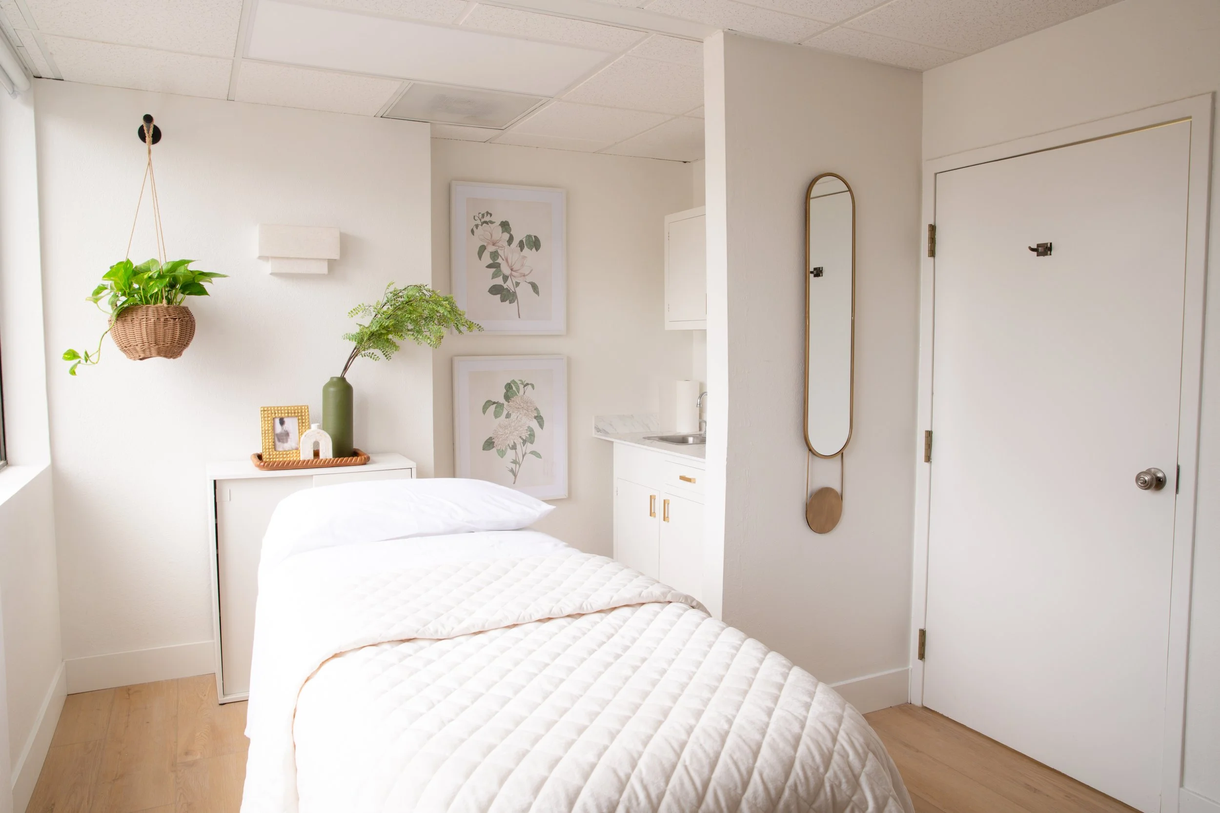 A cozy bedroom with a white bed, two framed floral art pieces on the wall, a white cabinet with decorative items and plants, a mirror on the wall, and a small kitchenette with a sink, all designed in a minimalist modern style.