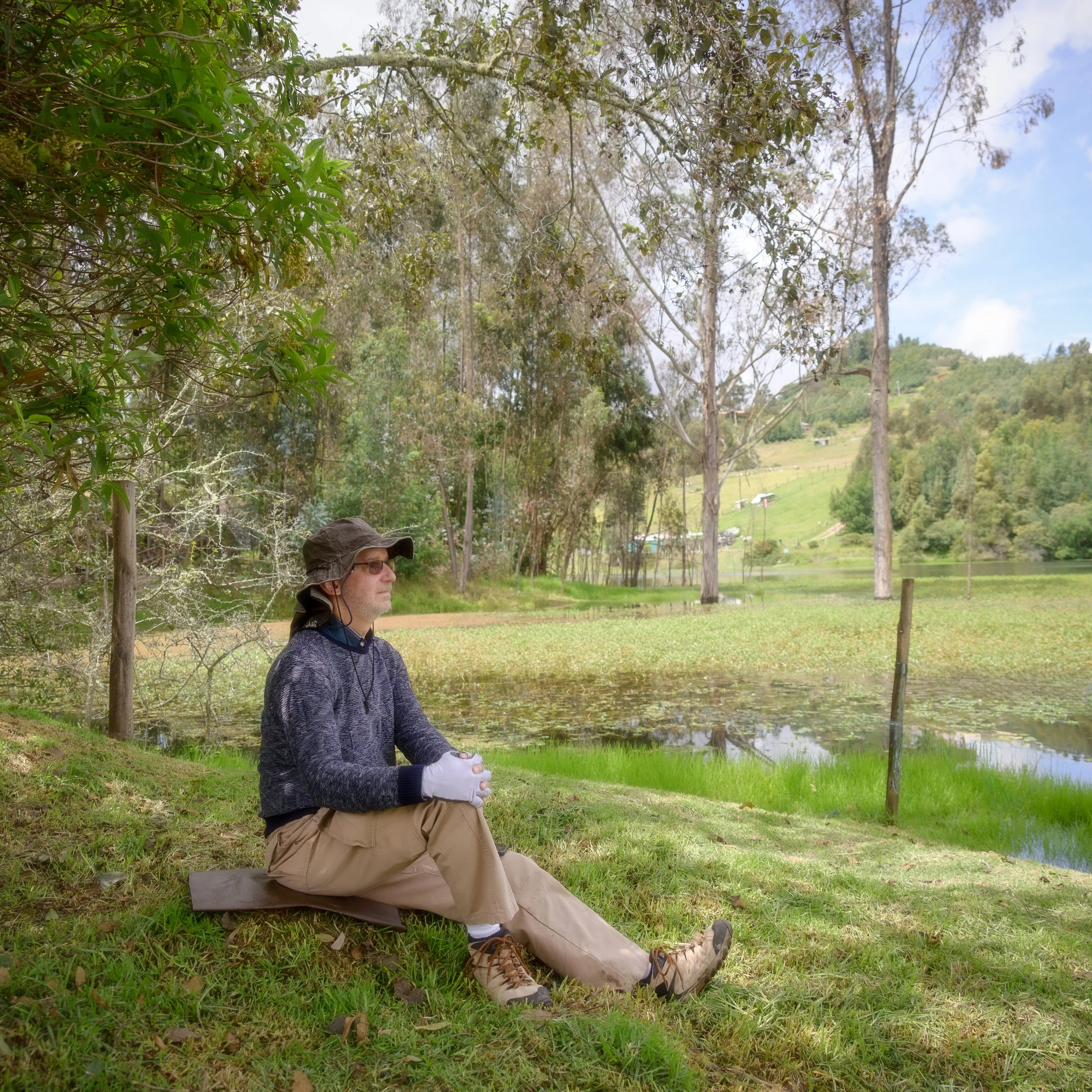 Luis Gerardo Alfaro sitting on a grass bank beside a pond, surrounded by trees and hills, wearing outdoor clothing and a hat, appearing relaxed and contemplative.